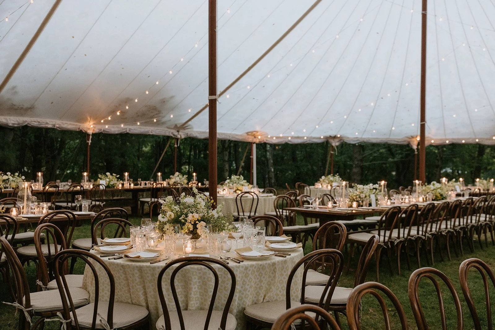 Outdoor event tent with round and long tables decorated with flowers and candles, surrounded by wooden chairs, set for a celebration at night.