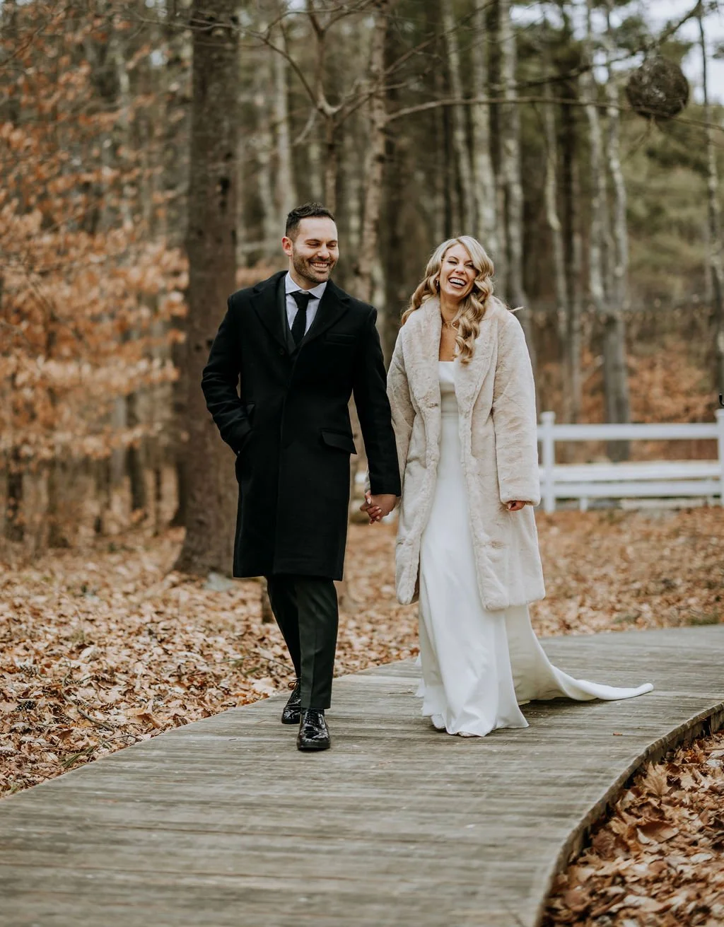 A happy couple in wedding attire holding hands and walking outdoors on a wooden path surrounded by autumn trees with leaves on the ground, smiling and looking joyful.