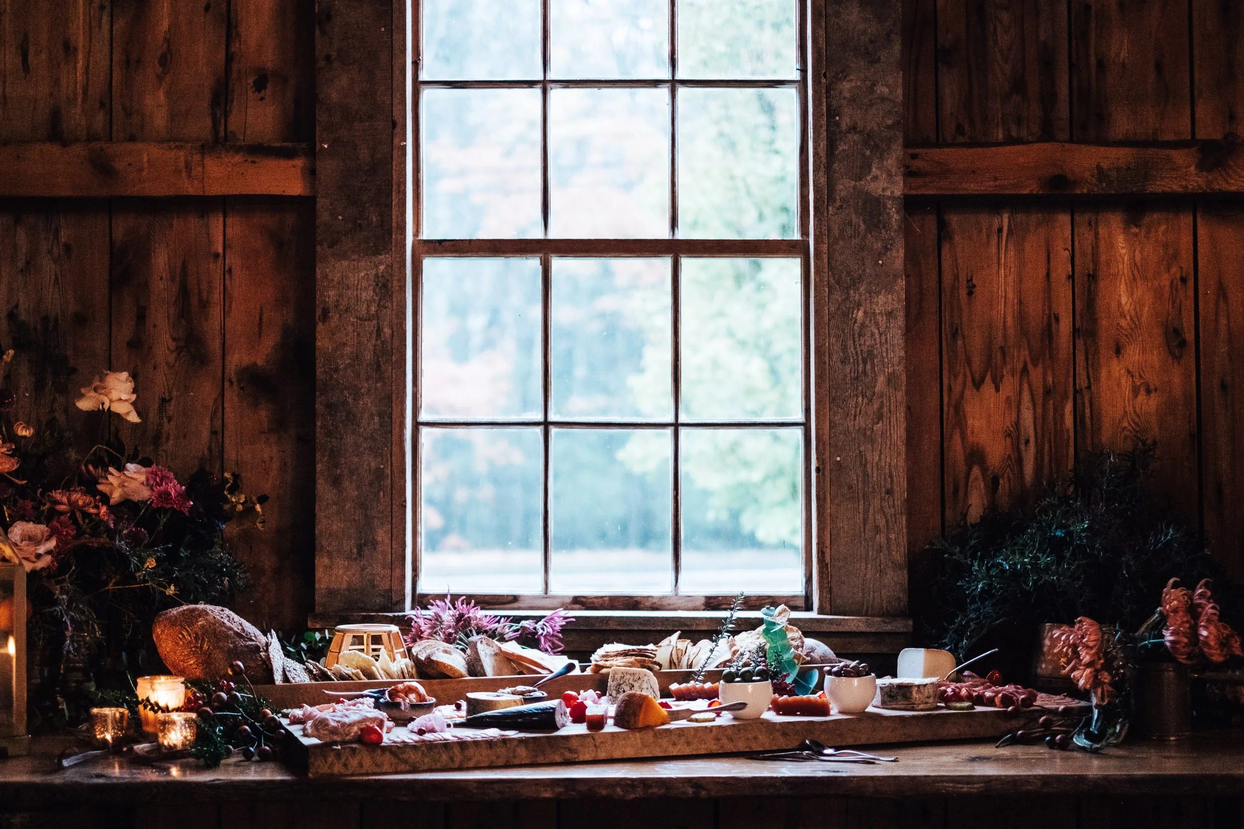 A rustic kitchen with a wooden countertop and a large window, displaying various cheeses, bread, fruits, and flowers on the counter, with greenery visible outside.
