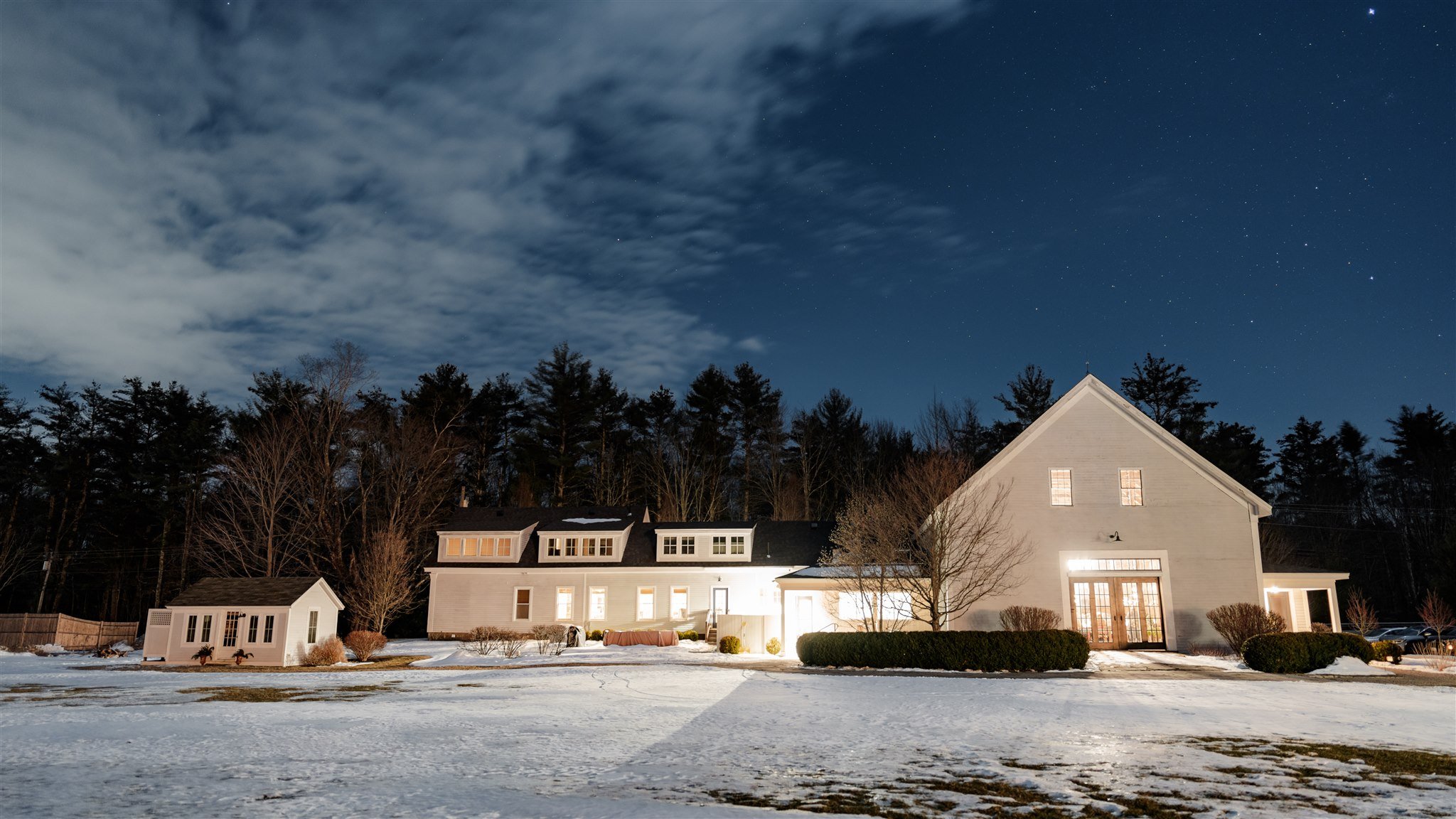 A large house with bright lights in a snowy yard at night, with trees and a partly cloudy sky overhead.