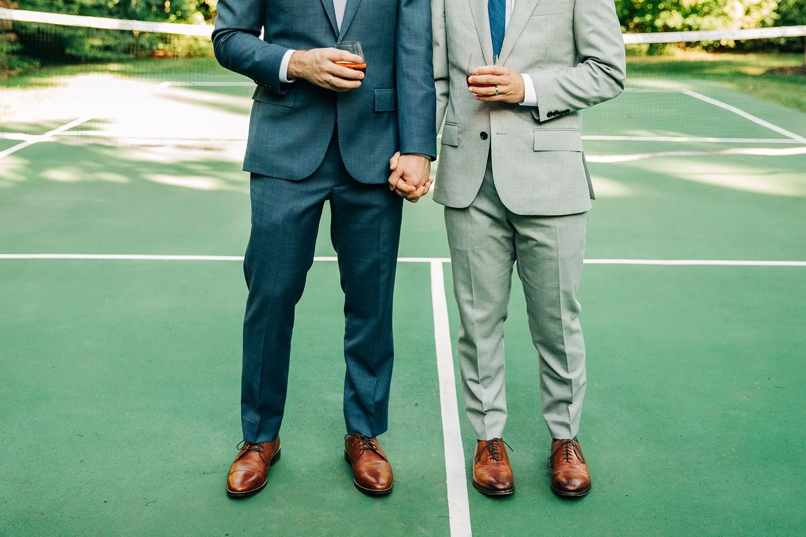 Two men in gray suits holding hands and drinks, standing on a green tennis court.