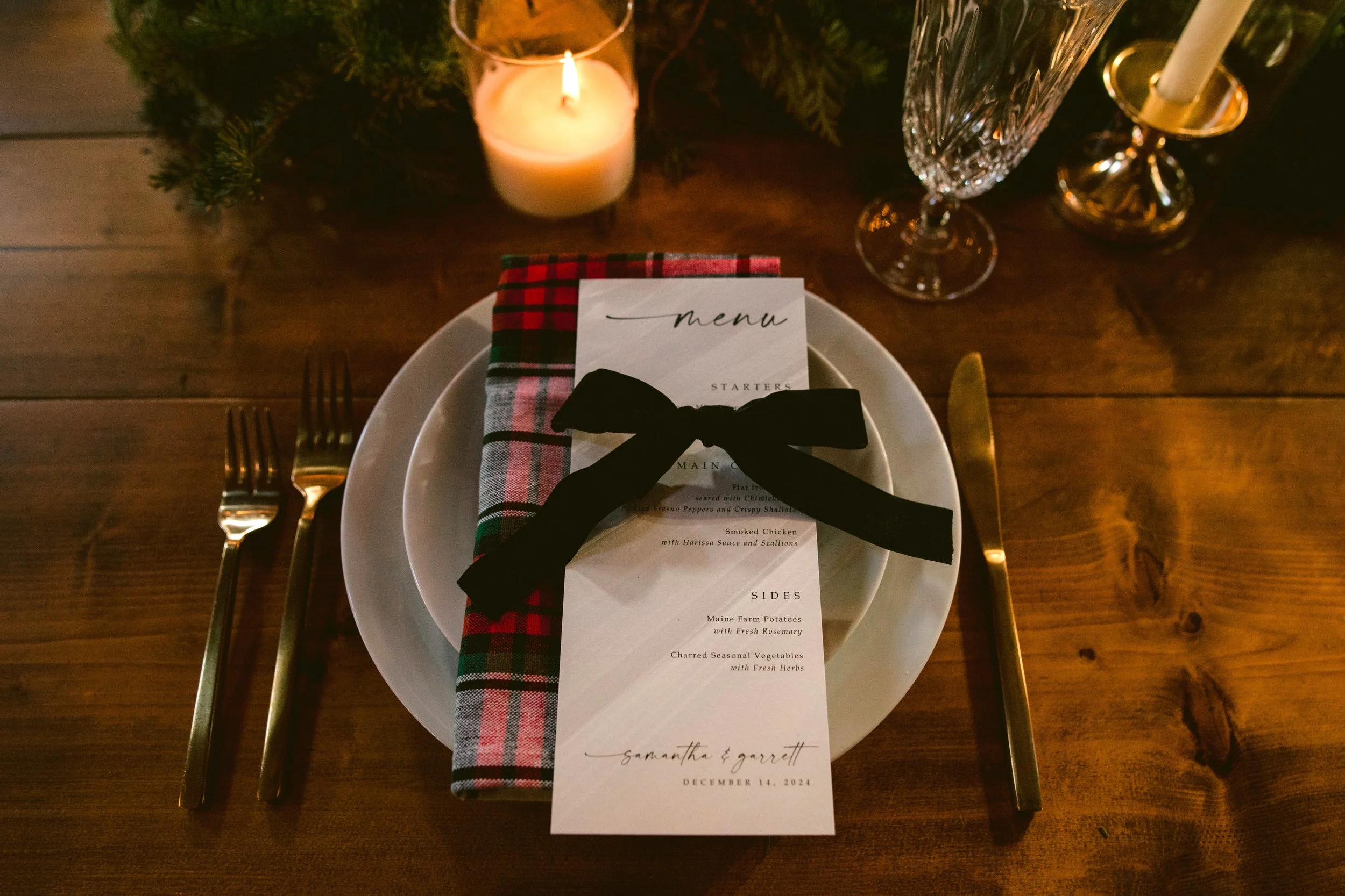 Festive table setting with a white plate, gold cutlery, a plaid napkin, a menu card tied with a black ribbon, a lit candle, and two wine glasses on a wooden table.