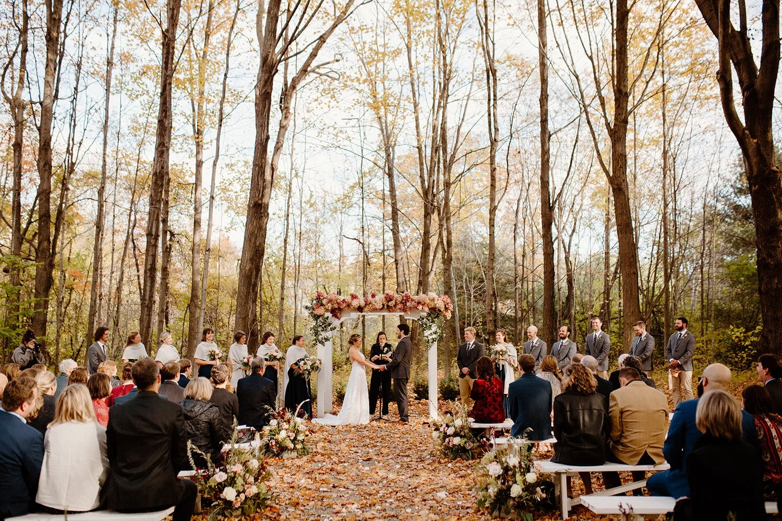 A wedding ceremony taking place outdoors in a wooded area with tall trees and fall foliage. The bride and groom are standing under a floral arch, exchanging vows, surrounded by wedding party and seated guests.