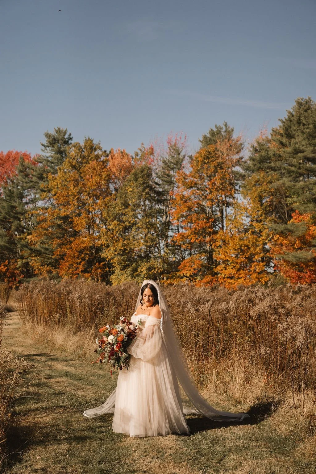 A bride in a cream-colored wedding dress with off-the-shoulder sleeves holding a large bouquet of red, orange, and white flowers stands on a grassy path surrounded by autumn trees with orange, yellow, and green leaves, under a clear blue sky.