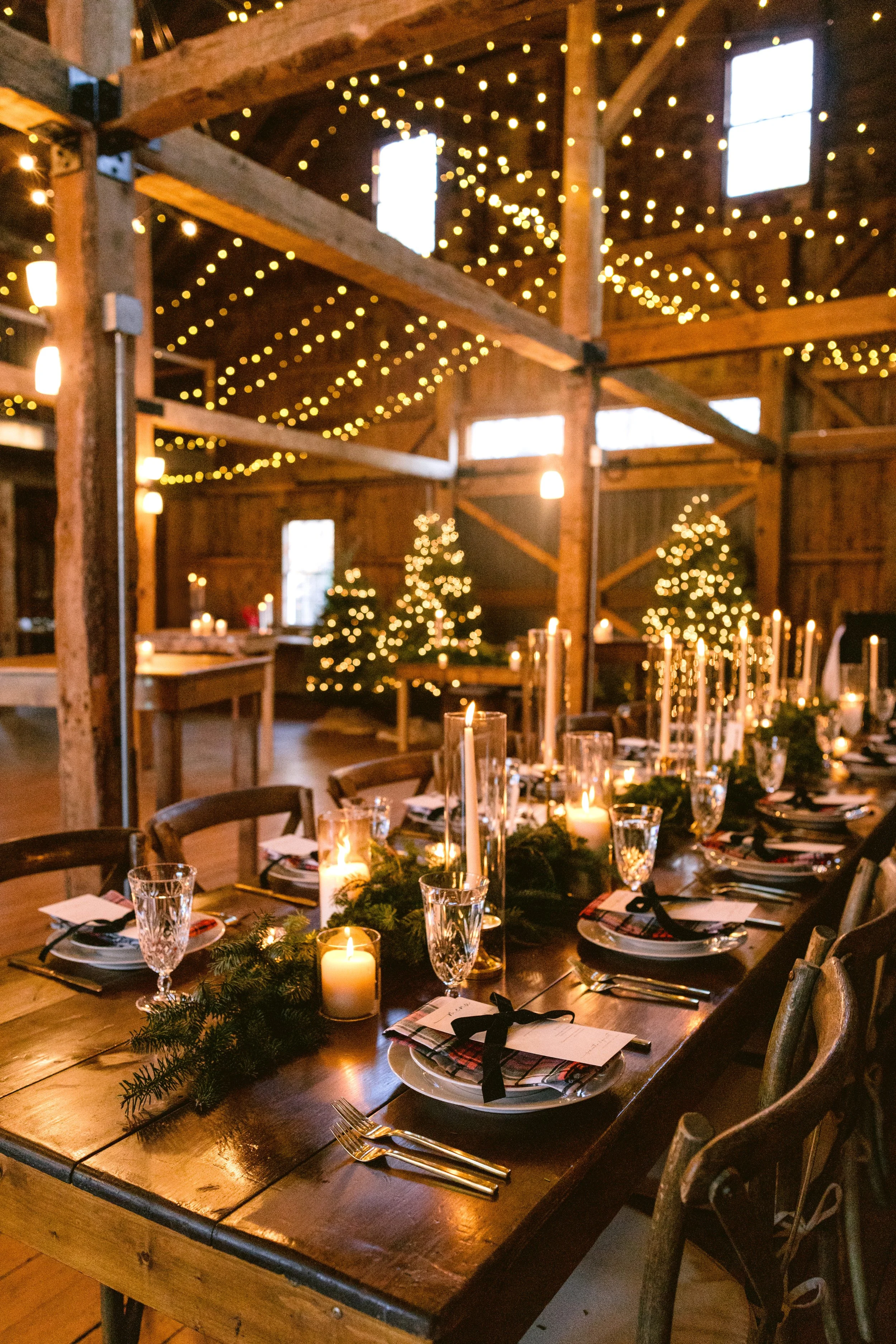 Rustic barn with wooden beams and string lights, decorated with small Christmas trees and a long dining table set for a holiday celebration with candles, wine glasses, and place settings.