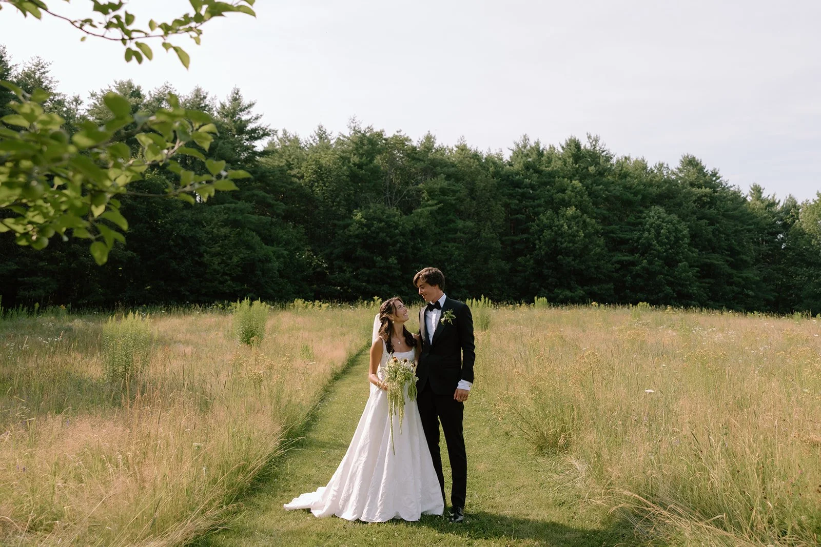 A bride and groom standing on a grassy path in a field with tall grass and wildflowers, surrounded by trees in the background, during a wedding photoshoot.