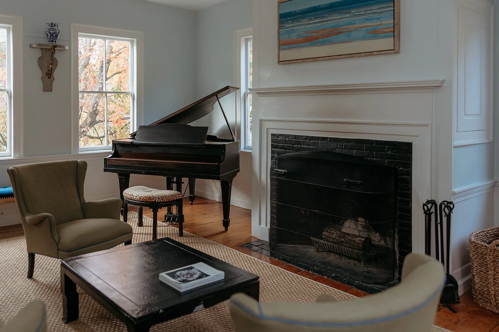 Living room with a black grand piano near a window, beige armchairs, a coffee table with a book, a fireplace with a white mantel, and artwork above the fireplace, in a home with hardwood floors and decorated walls.