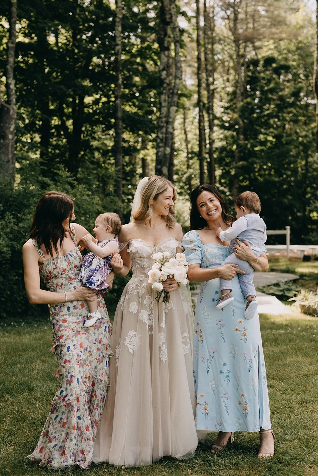 Four women and two children standing outdoors in a grassy area surrounded by tall trees, smiling and interacting during a sunny day. One woman is dressed in a wedding gown holding a bouquet, indicating a wedding celebration.