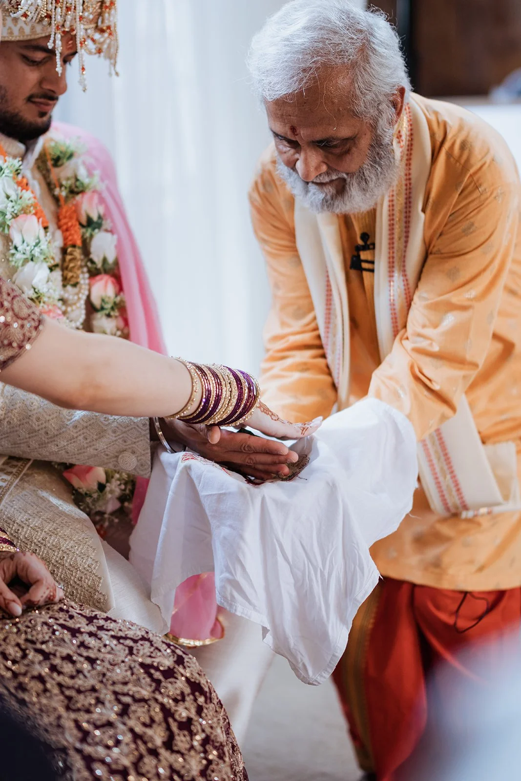 An elderly man performing a traditional Indian ceremony during a wedding, with a bride dressed in ornate attire and jewelry, and wearing red and gold bangles, while another man in traditional clothing looks on