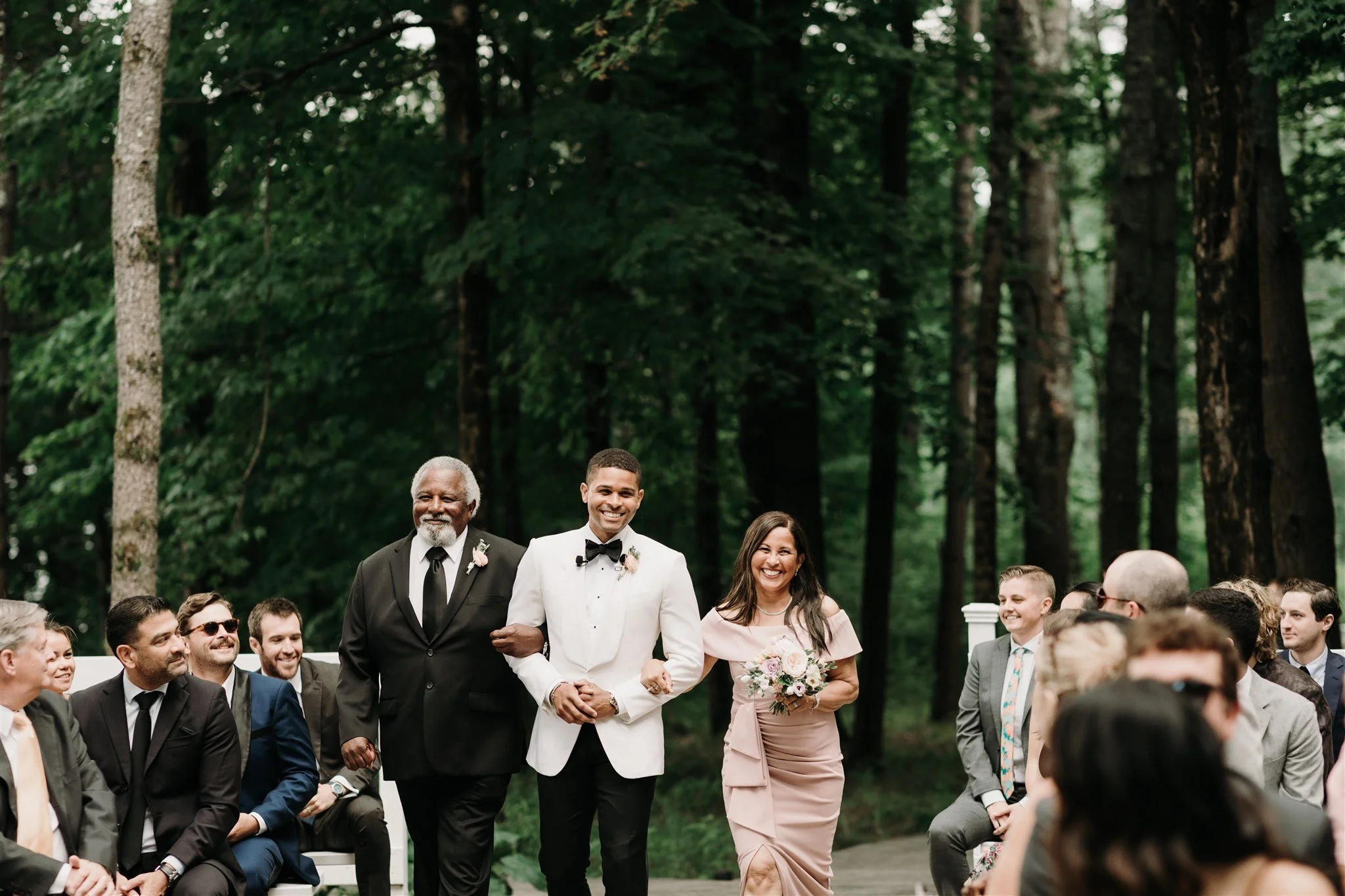 A wedding ceremony outdoors in a forest with three people walking down the aisle: a groom in a white tuxedo with black bow tie, an older man in a black suit, and a woman in a blush pink dress carrying a bouquet of flowers, surrounded by seated guests.