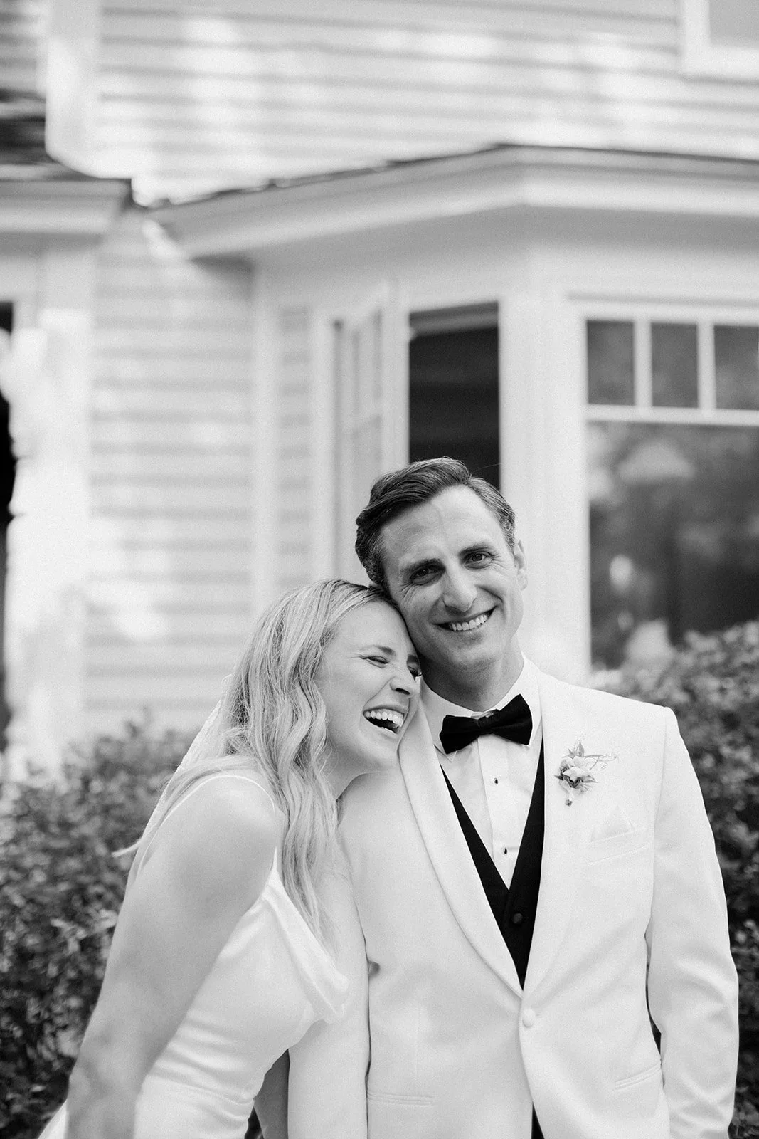 A black and white photo of a happy bride and groom, with the bride laughing and leaning her head on the groom's shoulder, standing outdoors in front of a house.