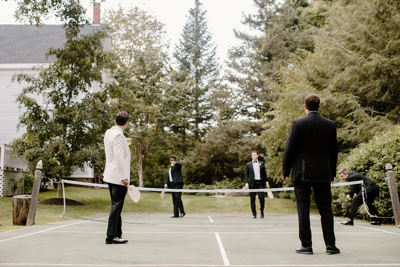 Five men playing badminton on an outdoor court, with trees and a house in the background. One man in a white tuxedo is holding a racket and standing on the left side. The other four men in black tuxedos are positioned around the net, with one pointing towards the court.