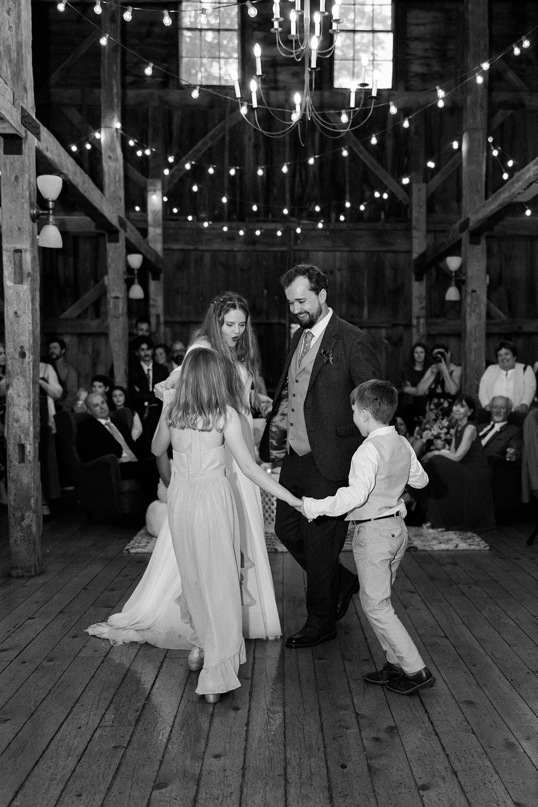 Black and white photo of a wedding reception in a rustic barn, with a bride, a groom, and two children dancing together surrounded by seated guests.