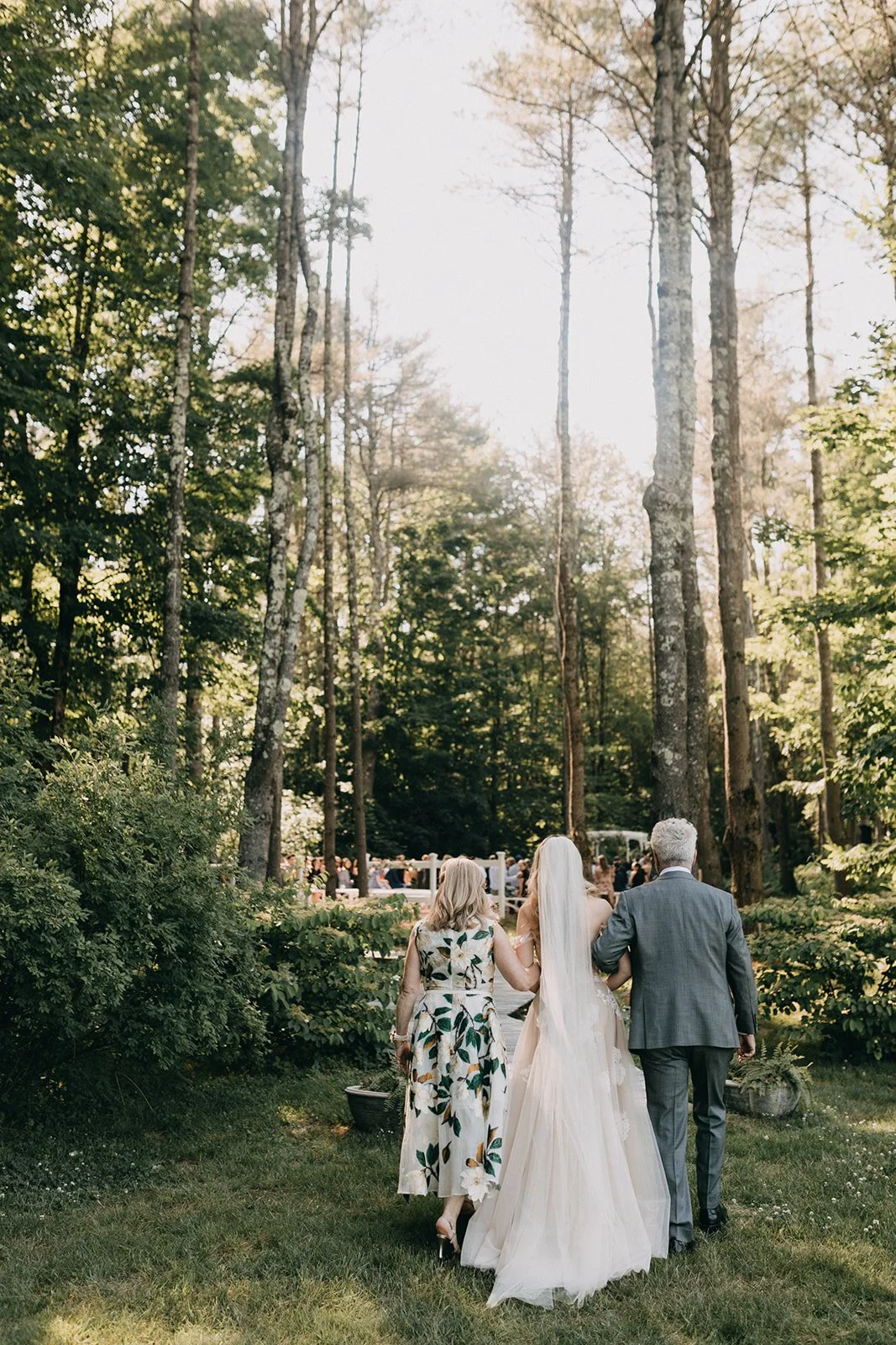 A bride in a wedding gown walking outdoors in a wooded area, accompanied by an older woman and a man, with ceremony attendees in the background.