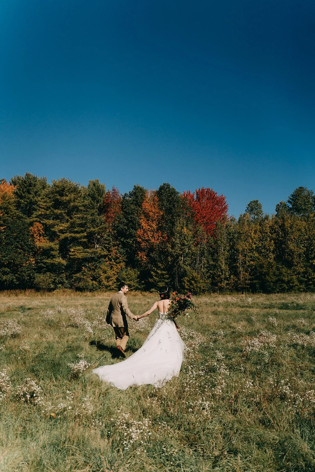 A bride and groom holding hands in a grassy field with fall foliage and trees in the background.