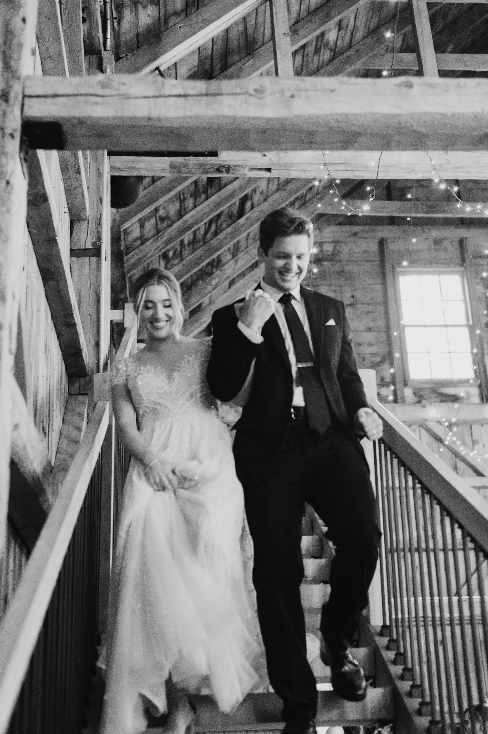 A black and white photo of a bride and groom celebrating on a wooden staircase in a rustic barn, smiling and dancing with fairy lights in the background.