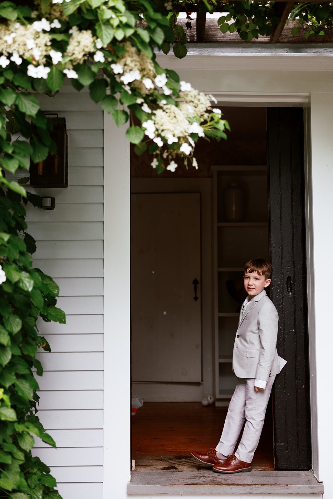 A young boy in a light gray suit and brown shoes standing at a doorway of a house, with greenery and white flowers hanging above him.
