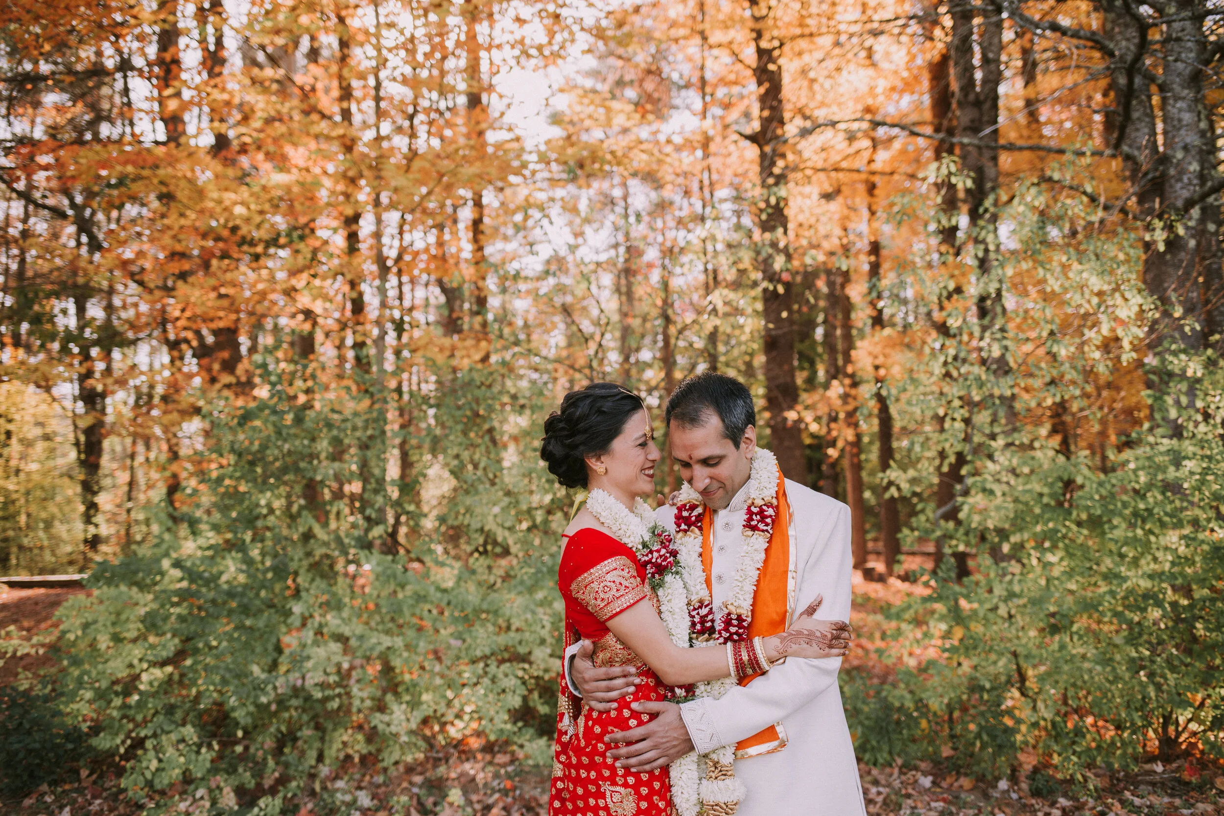 A couple dressed in traditional Indian wedding attire sharing an embrace in a forest during autumn.