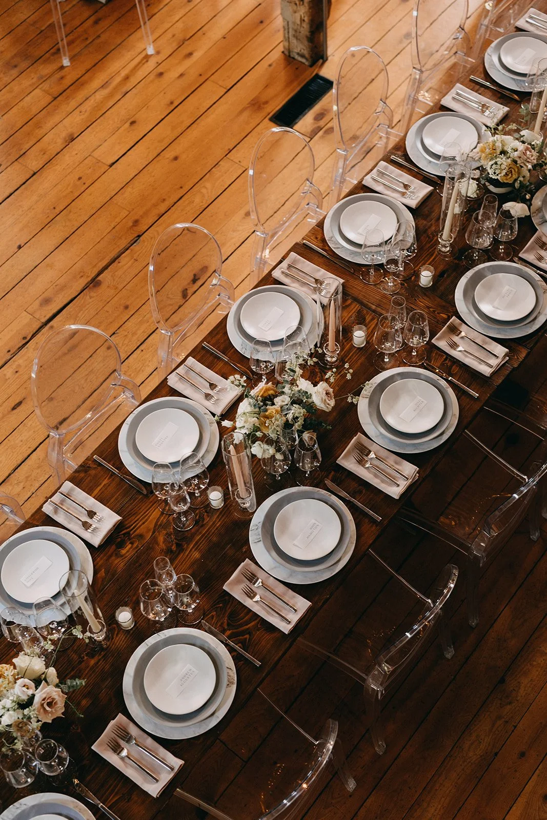 A long wooden dining table set for a formal event with white and gray tableware, clear acrylic chairs, floral centerpieces, and candles, on a wooden floor.