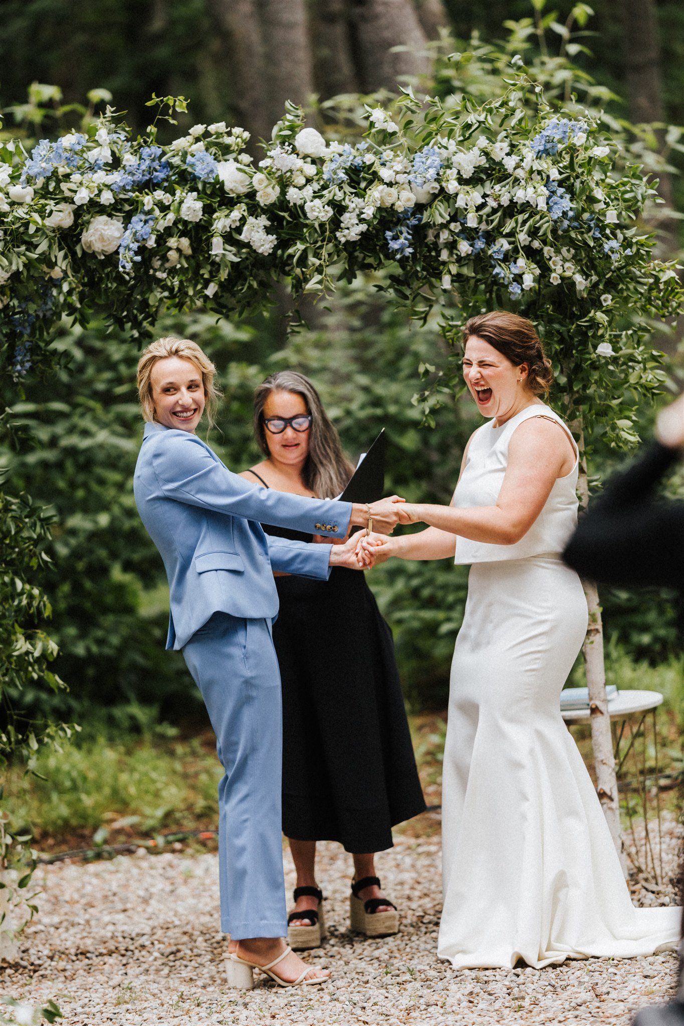Two women holding hands and smiling at each other during a wedding ceremony under a floral archway in a forest setting, with a woman officiant standing behind them.