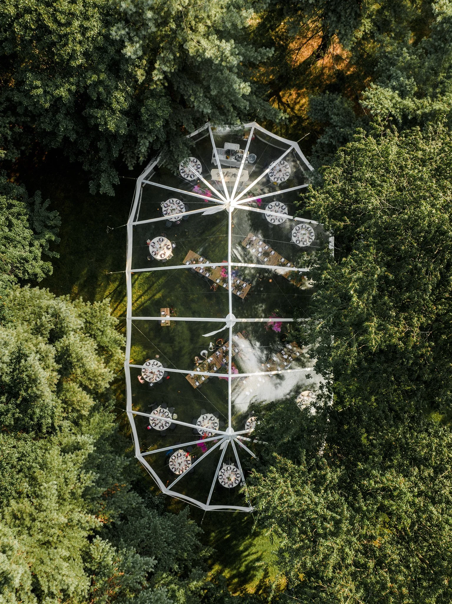 Aerial view of an outdoor event with tables set for dining, surrounded by dense green trees, with a large transparent tent covering the area.