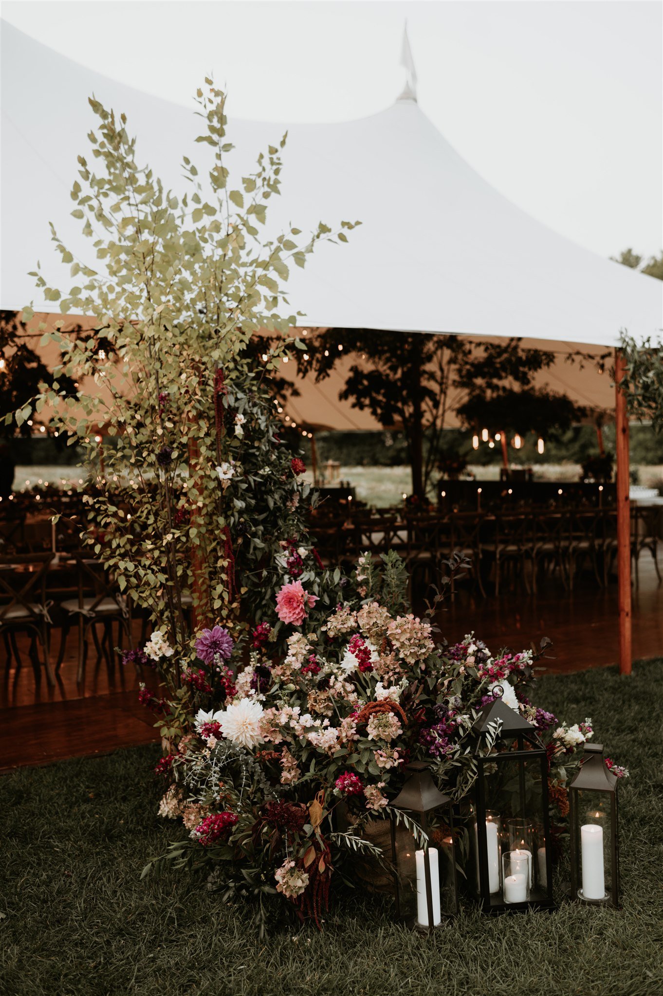Decorative floral display with candles in lanterns at an outdoor event under a white tent.