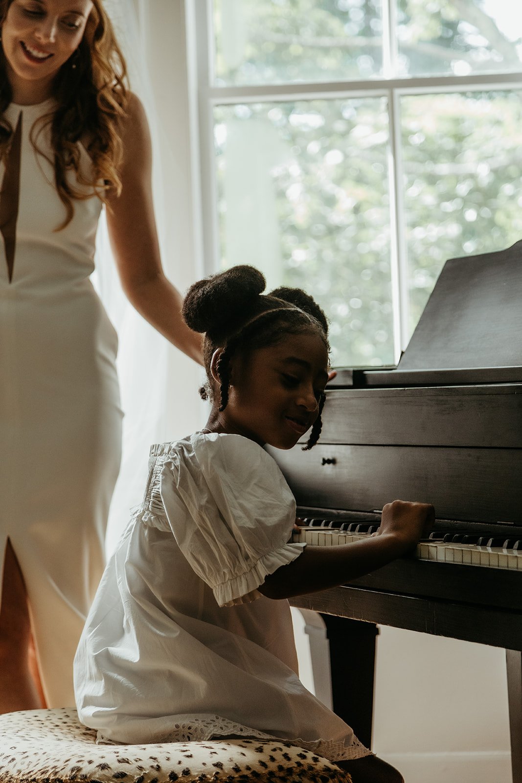 A young girl playing piano with an adult woman standing beside her, both inside a bright room with a window in the background.