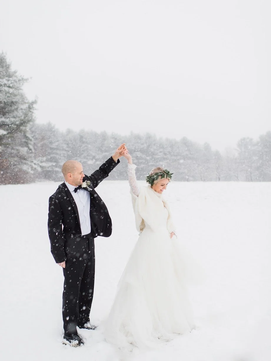 Newlywed couple dancing in a snowy landscape, the groom in a black tuxedo and the bride in a white wedding dress with a floral crown, holding hands and smiling.