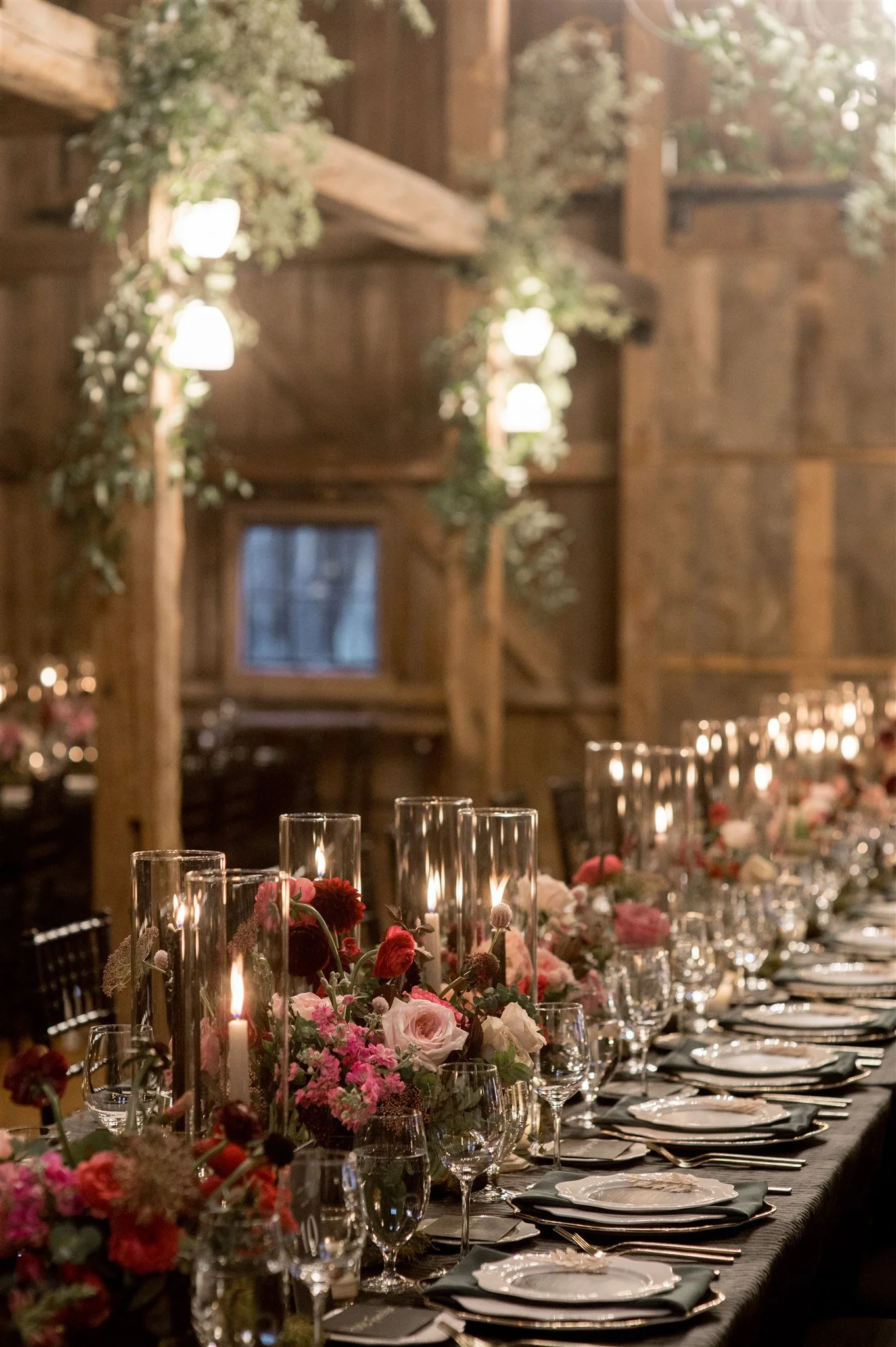 A long banquet table decorated with pink and red flowers, lit candles, and glassware in a rustic wooden venue.