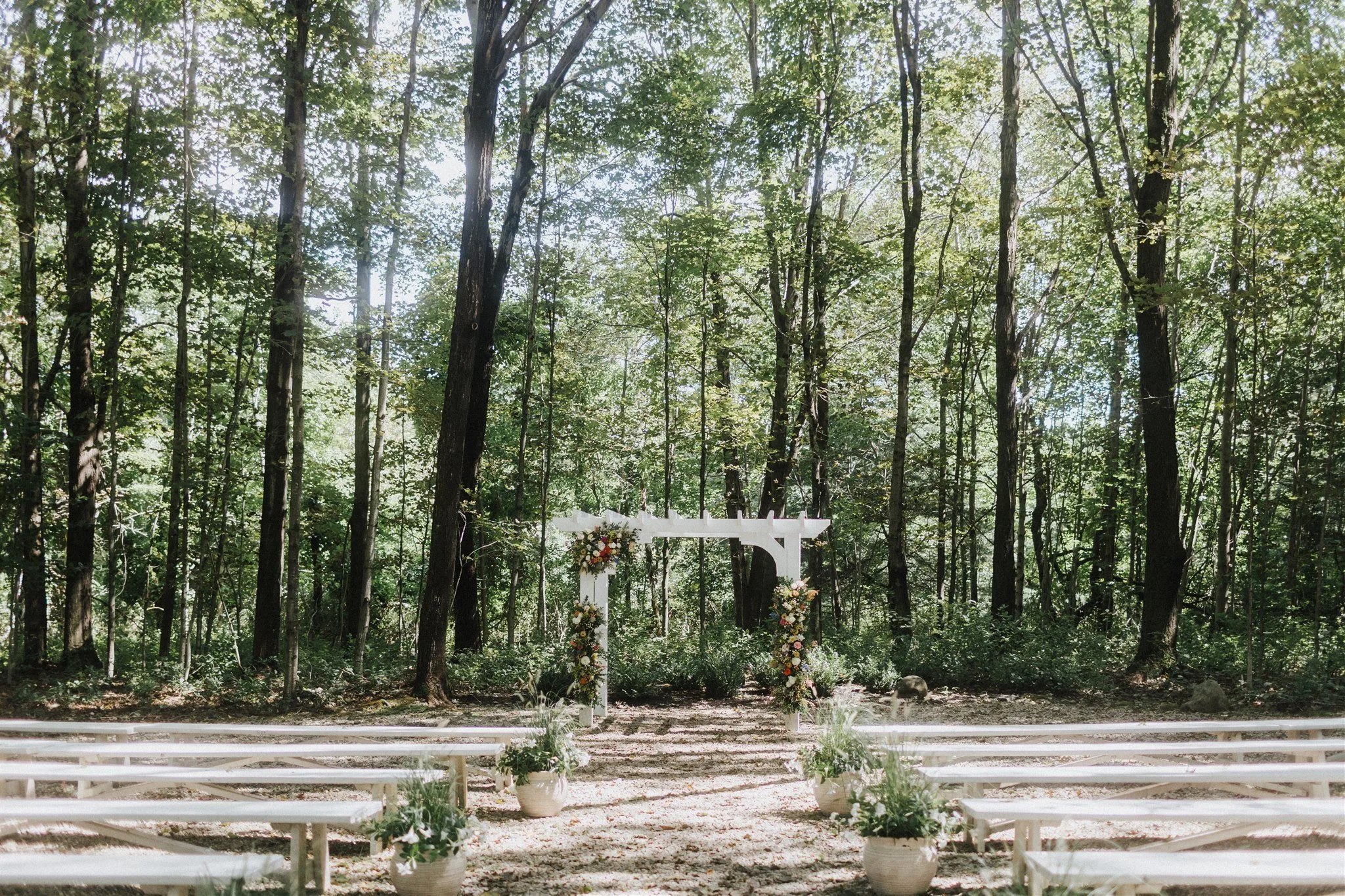 Outdoor wedding ceremony setup in a forest with white benches and floral arrangements on a white arch.
