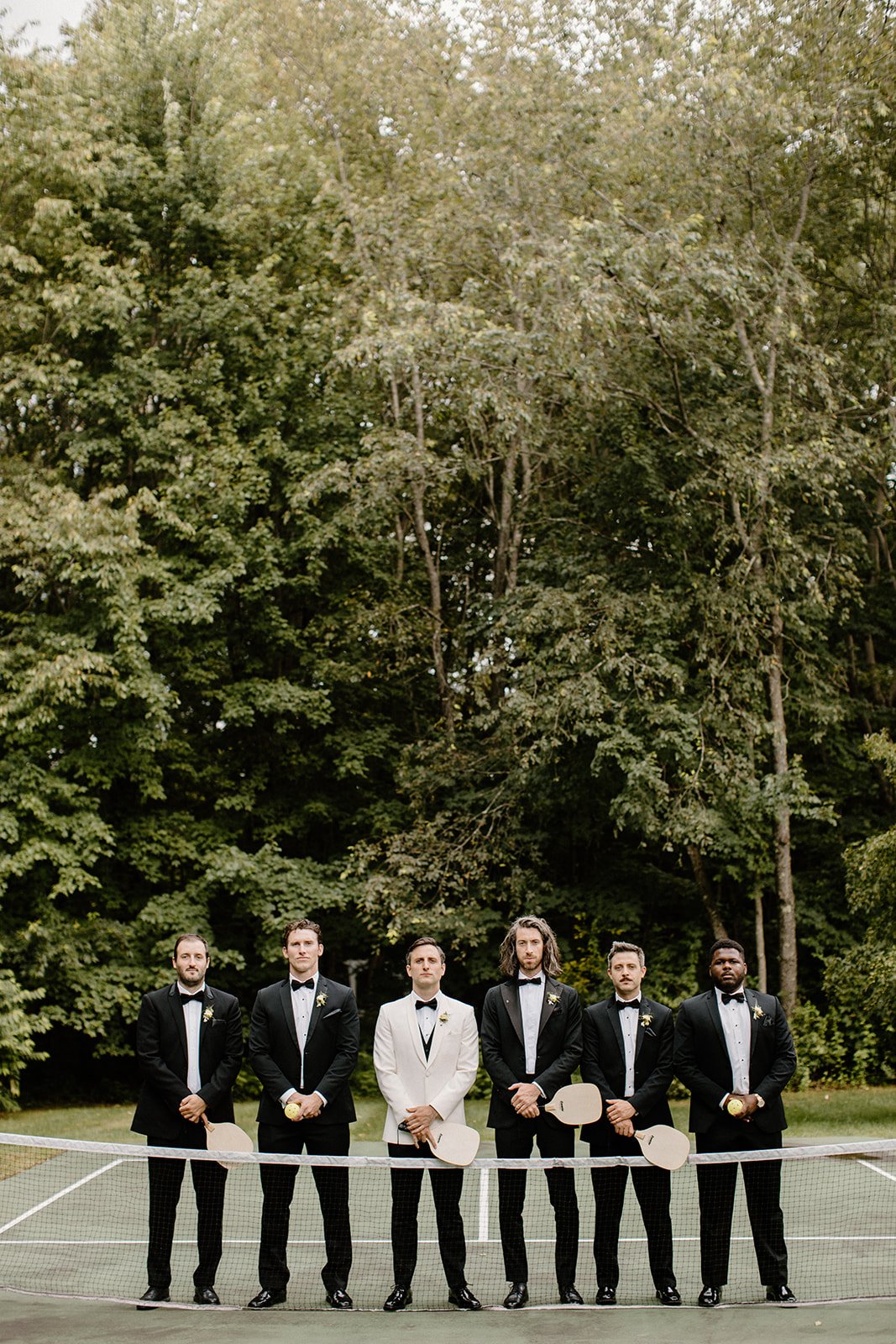 Six men dressed in tuxedos standing on a tennis court with a net in front of them and trees behind.