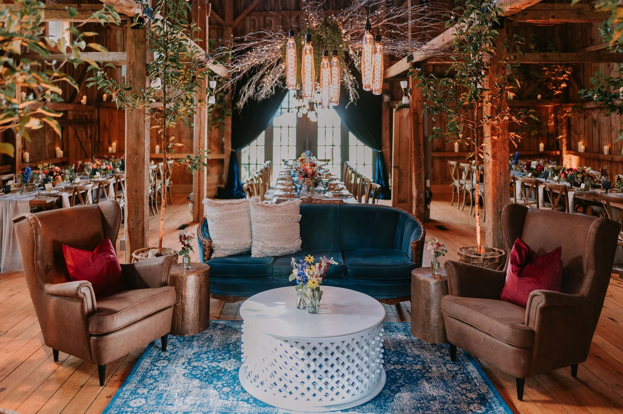 Interior of a rustic wedding or event venue with wood-paneled walls and ceiling, featuring a chandelier with multiple hanging light fixtures. In the foreground, there are two armchairs with pillows, a blue velvet loveseat, a round white coffee table,