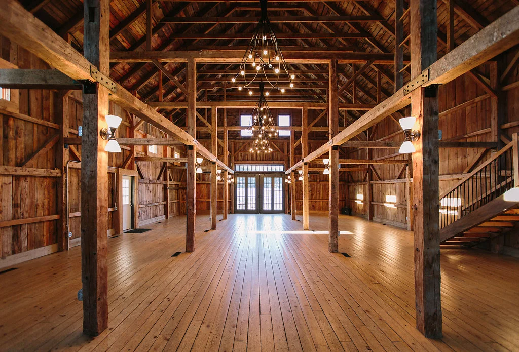 Interior of a large wooden barn with high vaulted ceiling, wooden beams, and a polished wooden floor. Chandeliers hang from the ceiling, and walls are lined with windows and wall-mounted lights. There is a staircase on the right side leading upstairs