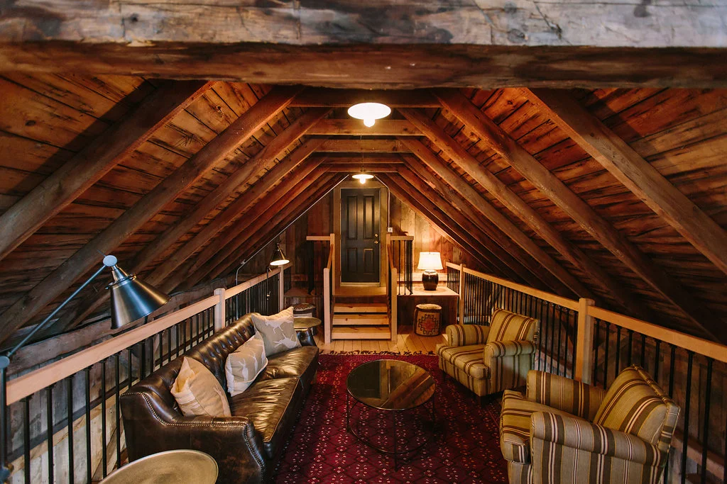 Cozy attic with wooden walls and ceiling, featuring a leather sofa, striped armchairs, a round coffee table, a lamp, and a door at the end.
