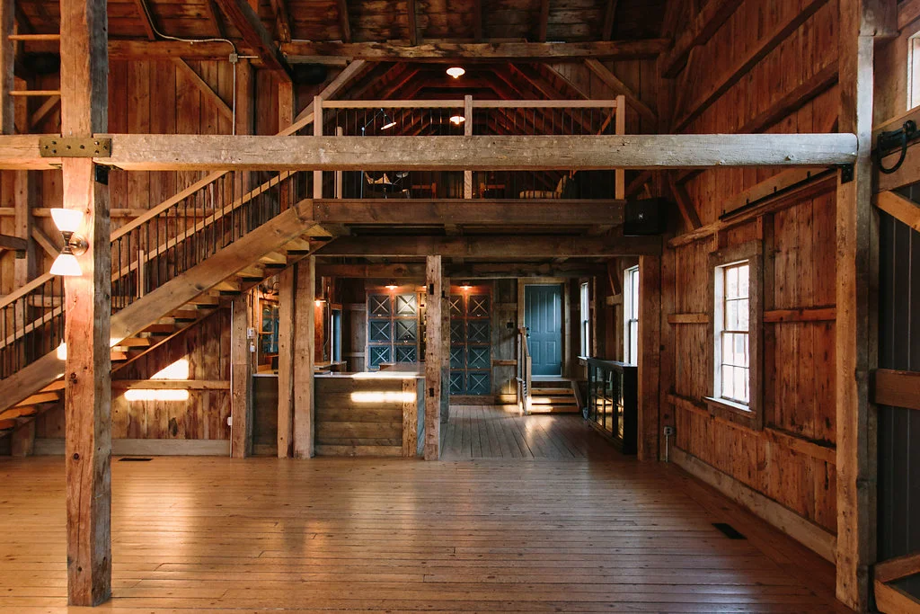 Interior of a rustic barn with wooden walls, exposed beams, and a staircase leading to a loft area. Natural light from windows illuminates the space, and there is a door at the back.