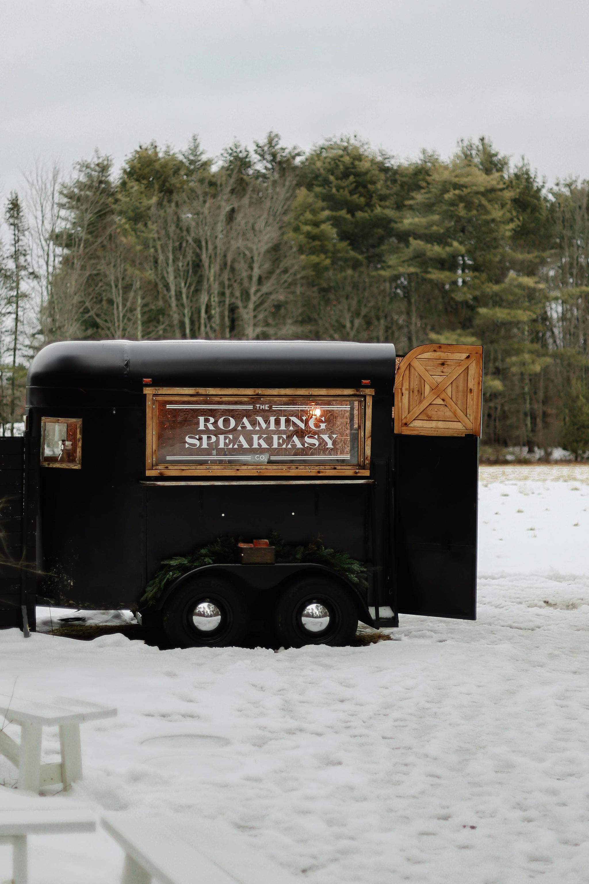 A black food truck called 'The Roaming Speakeasy' parked outdoors on snow-covered ground, with a wooded background.