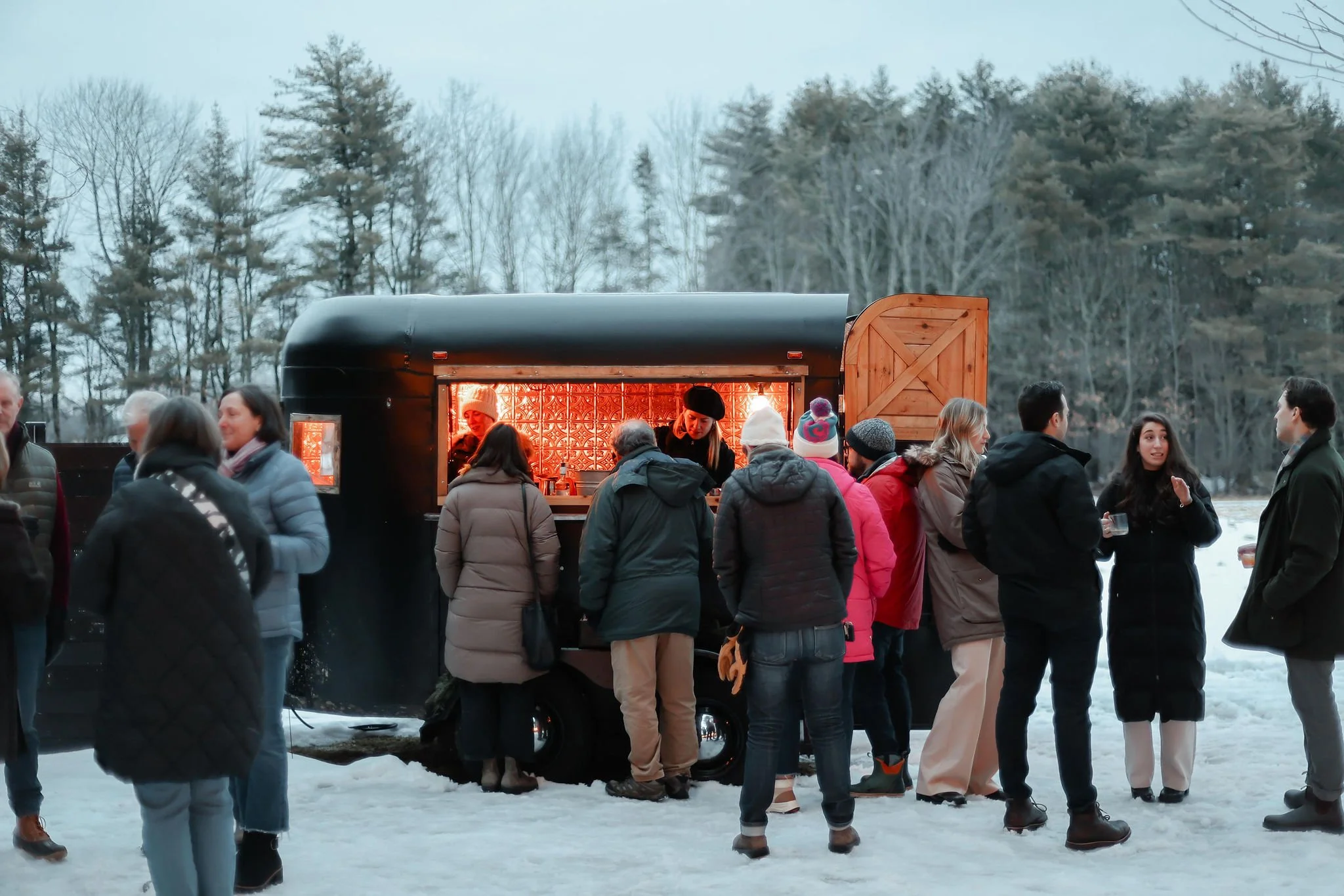People gathered around a mobile bar in a snowy outdoor setting with trees in the background.