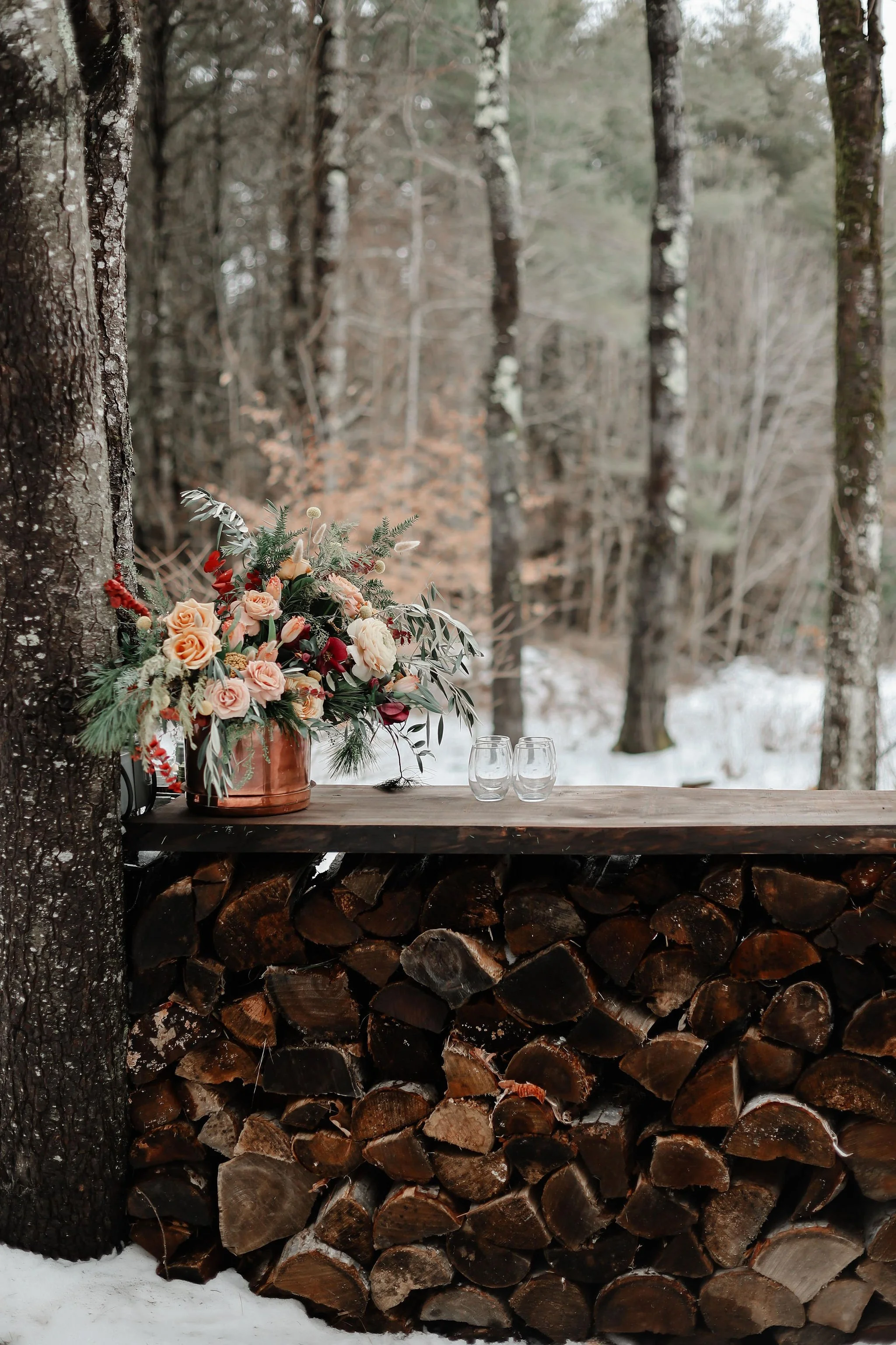 A rustic outdoor setting with a large bouquet of flowers in a copper vase and two empty glasses on a wooden table, in a snowy forest environment.