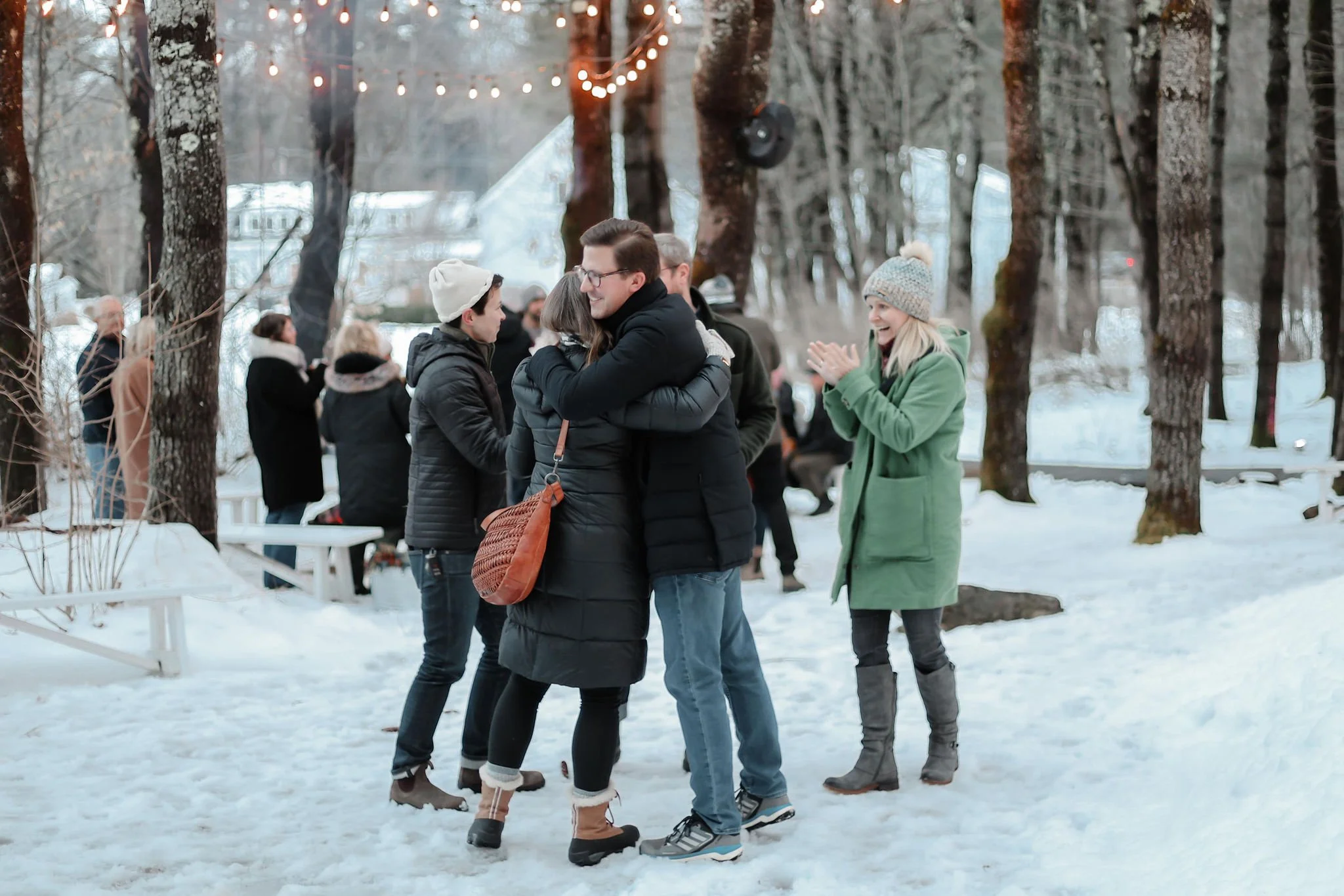 People in winter clothing hugging and smiling at a snowy outdoor gathering, with a woman clapping nearby.