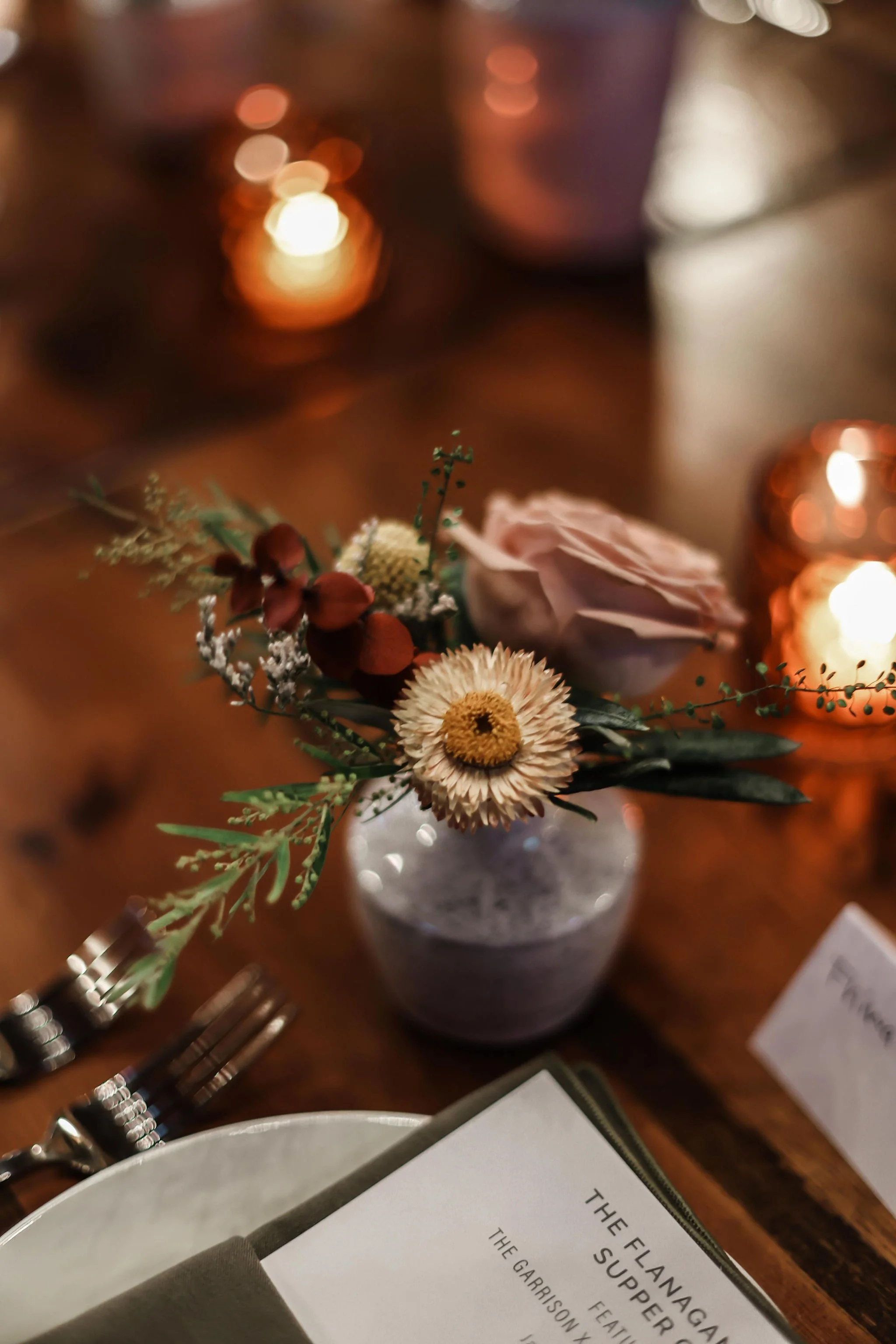 A small vase with a dried flower arrangement sitting on a wooden table, with candles and a menu nearby.
