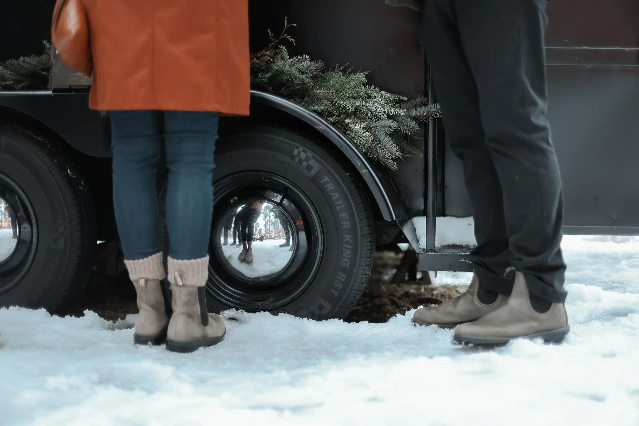 People standing in the snow near a black trailer truck decorated with evergreen branches.