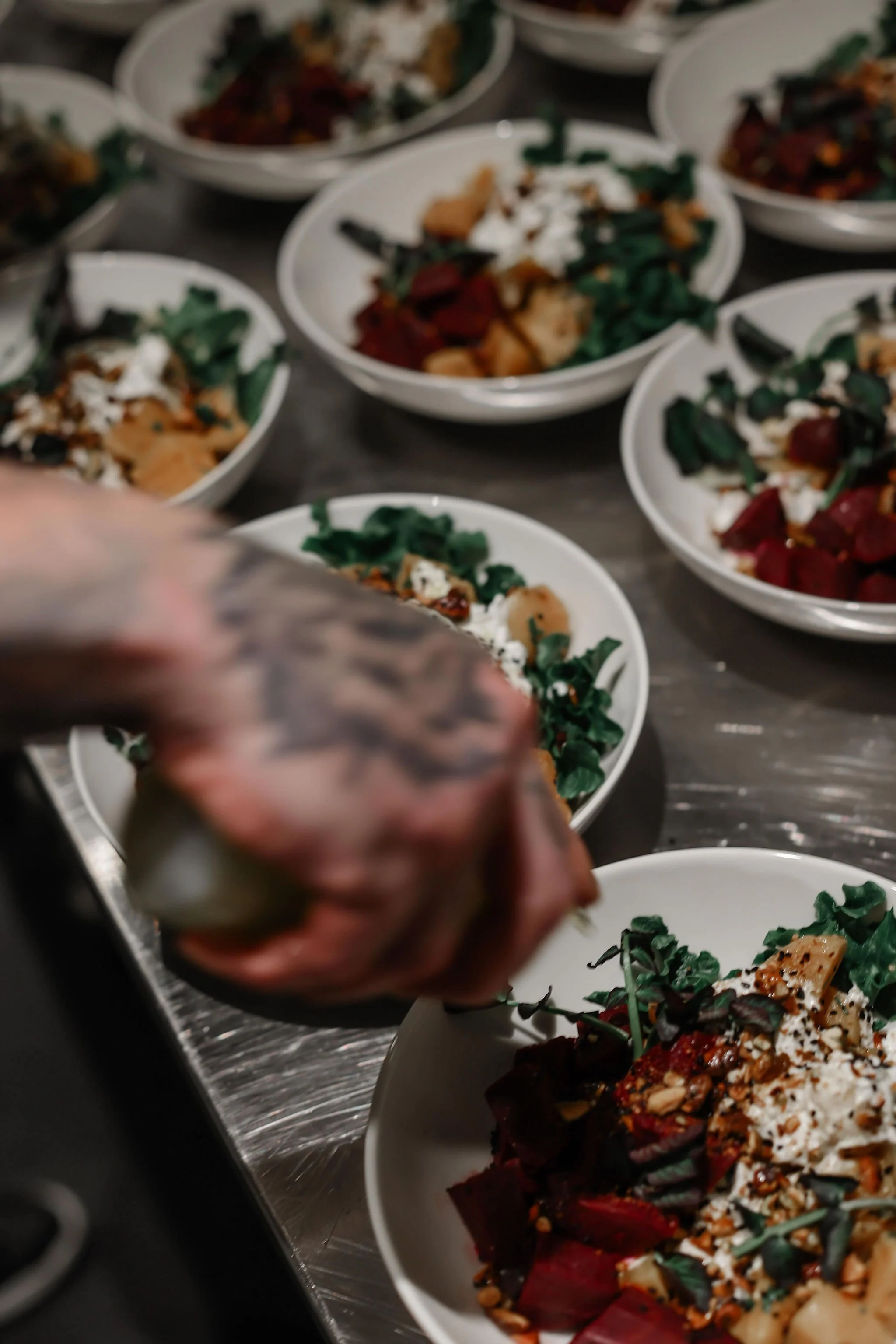 Chef garnishing bowls of salad with herbs on a stainless steel counter.
