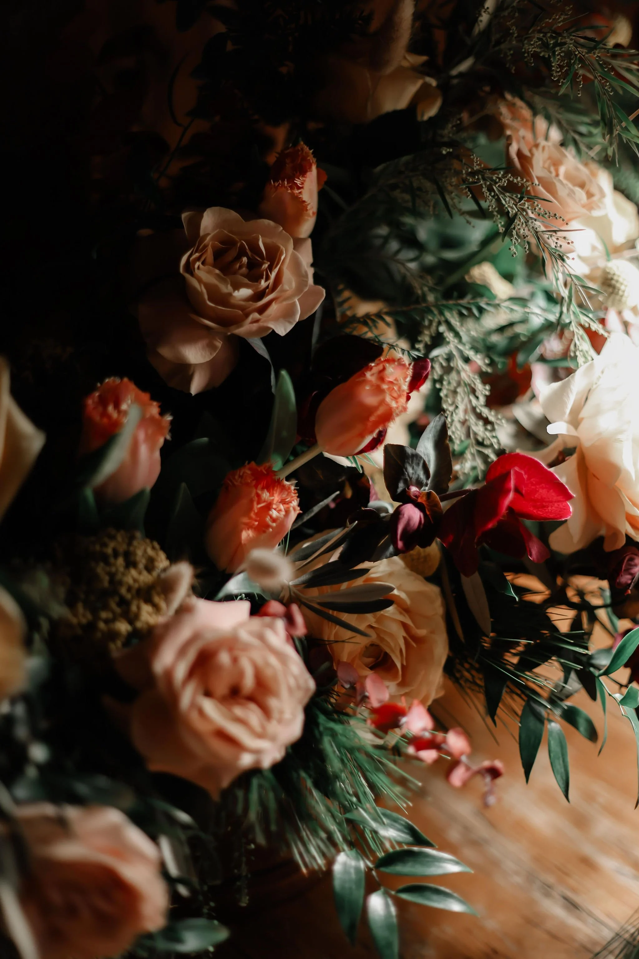 Close-up of a diverse array of peach, pink, and red flowers with green foliage, illuminated by warm light, arranged on a wooden surface.