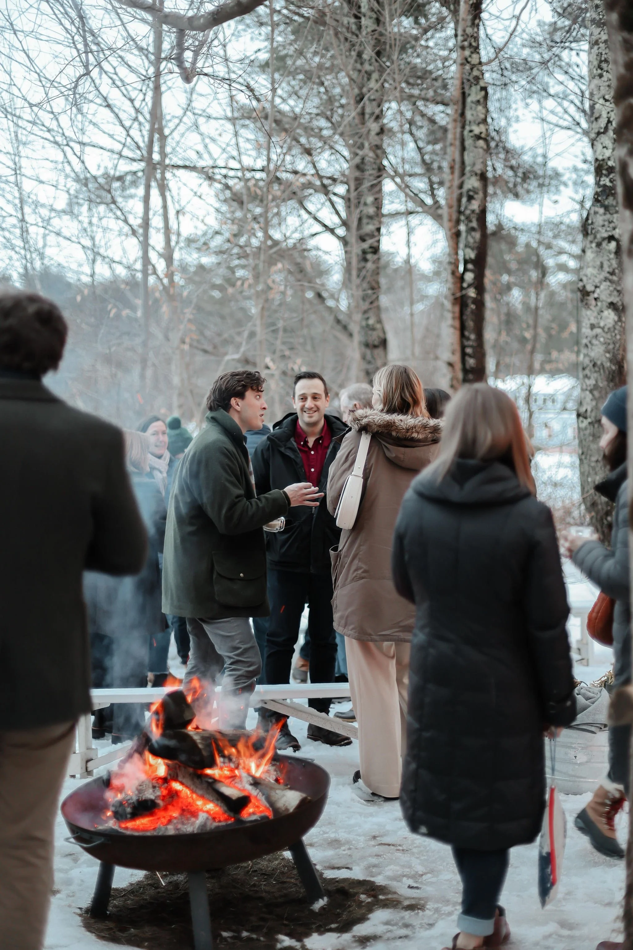 People gathered outdoors in winter, standing around a fire pit with snow on the ground, talking and socializing among trees.