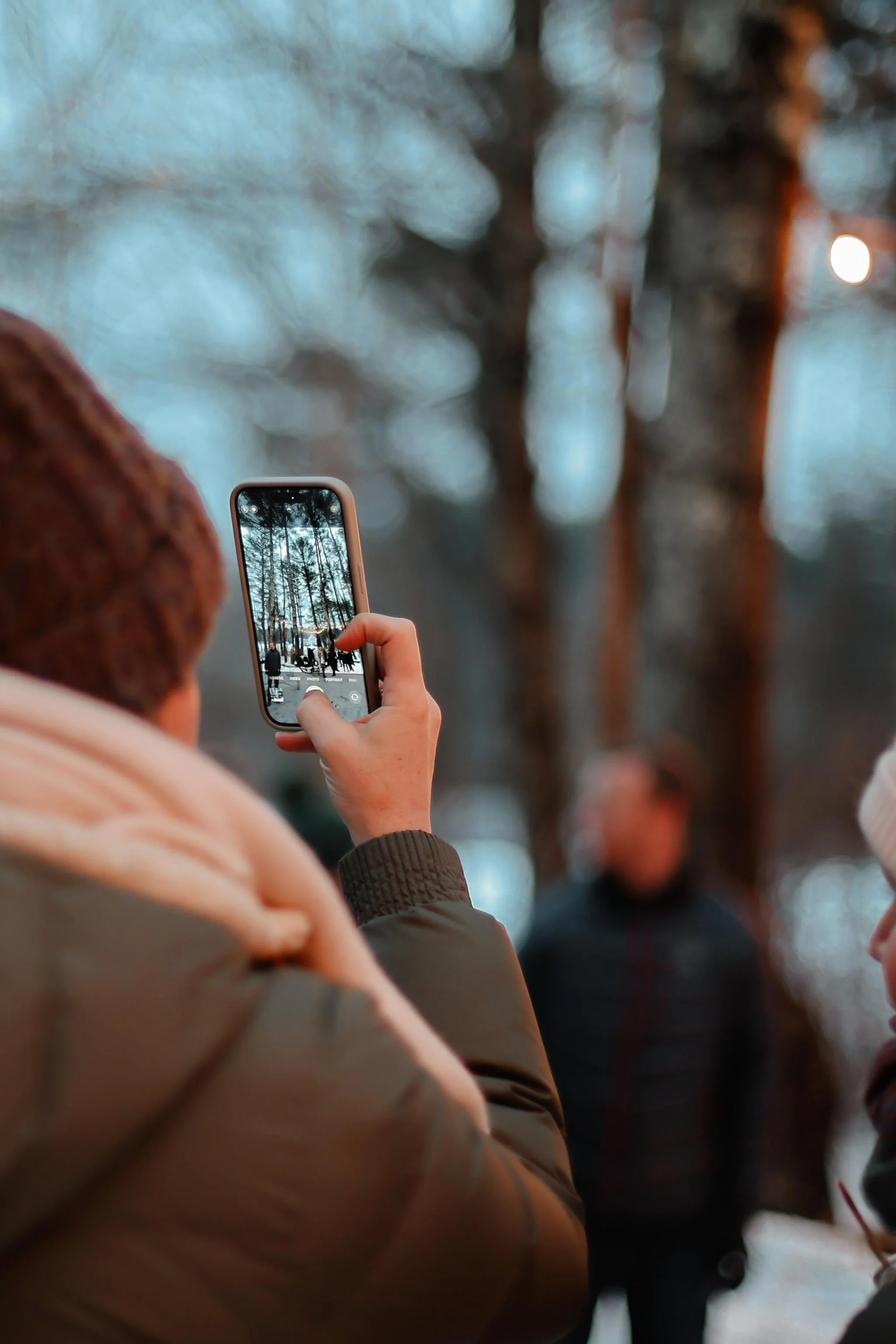 Person taking a photo with a smartphone in a wooded outdoor setting, with trees and other people in the background.