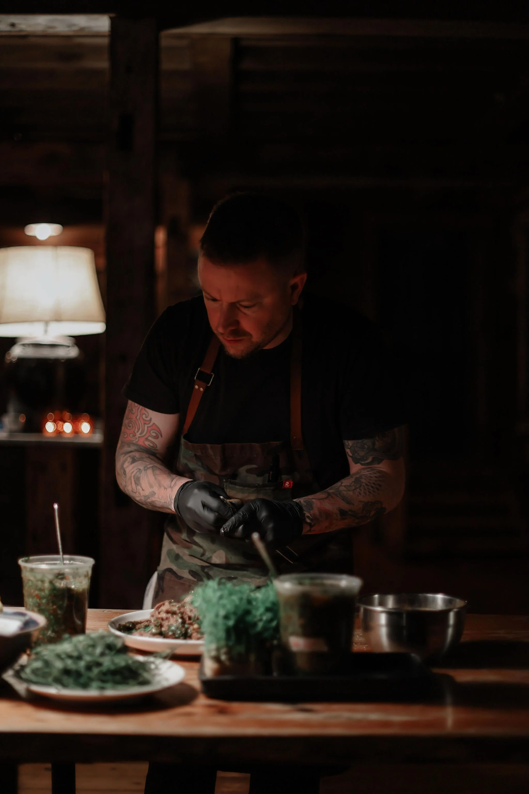 A man with tattoos wearing black gloves and a camouflage apron is preparing food in a dimly lit rustic kitchen, with plates of food and drinks on a wooden table.