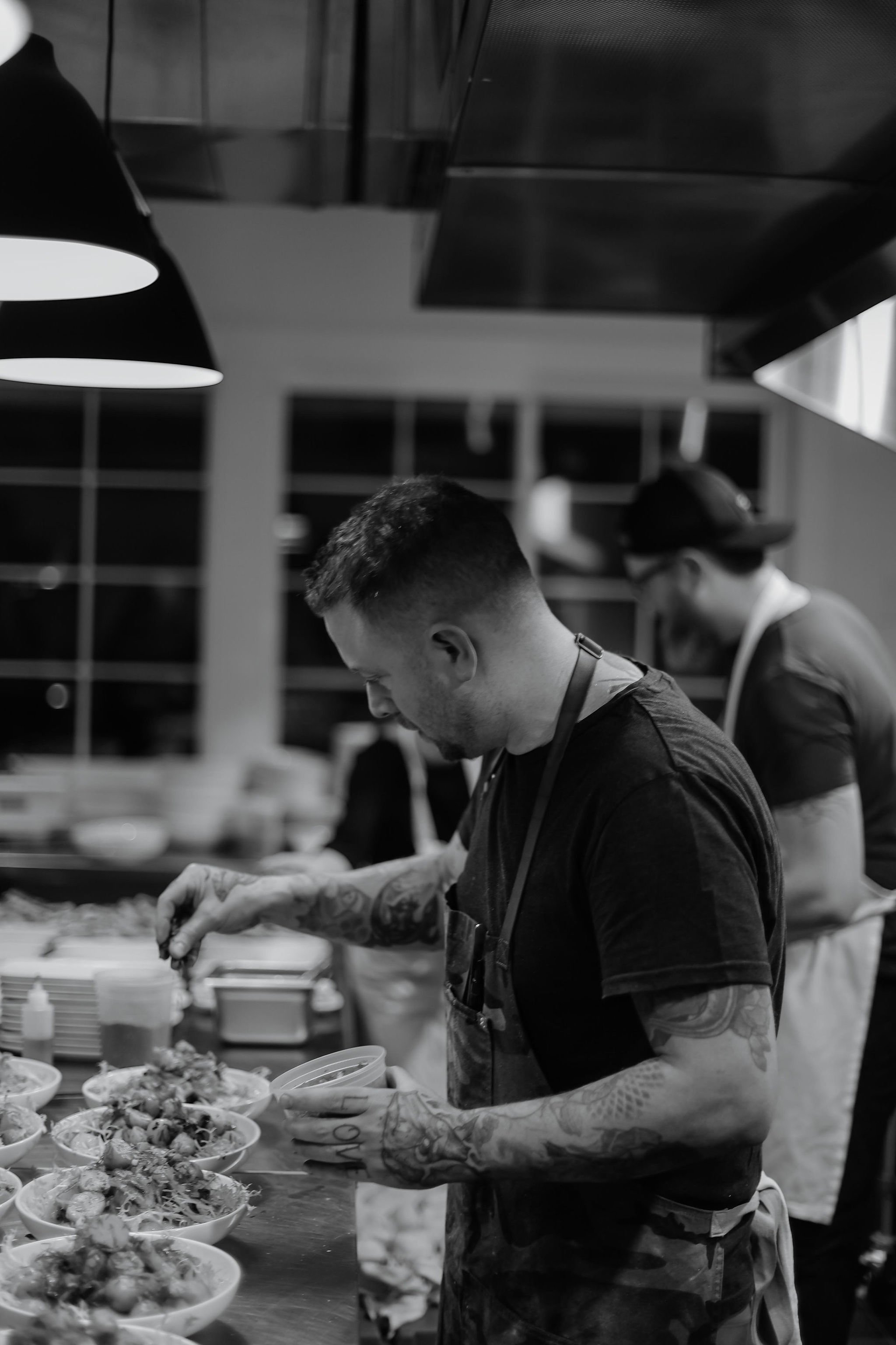 A man with tattoos preparing food in a professional kitchen, with another person in the background.