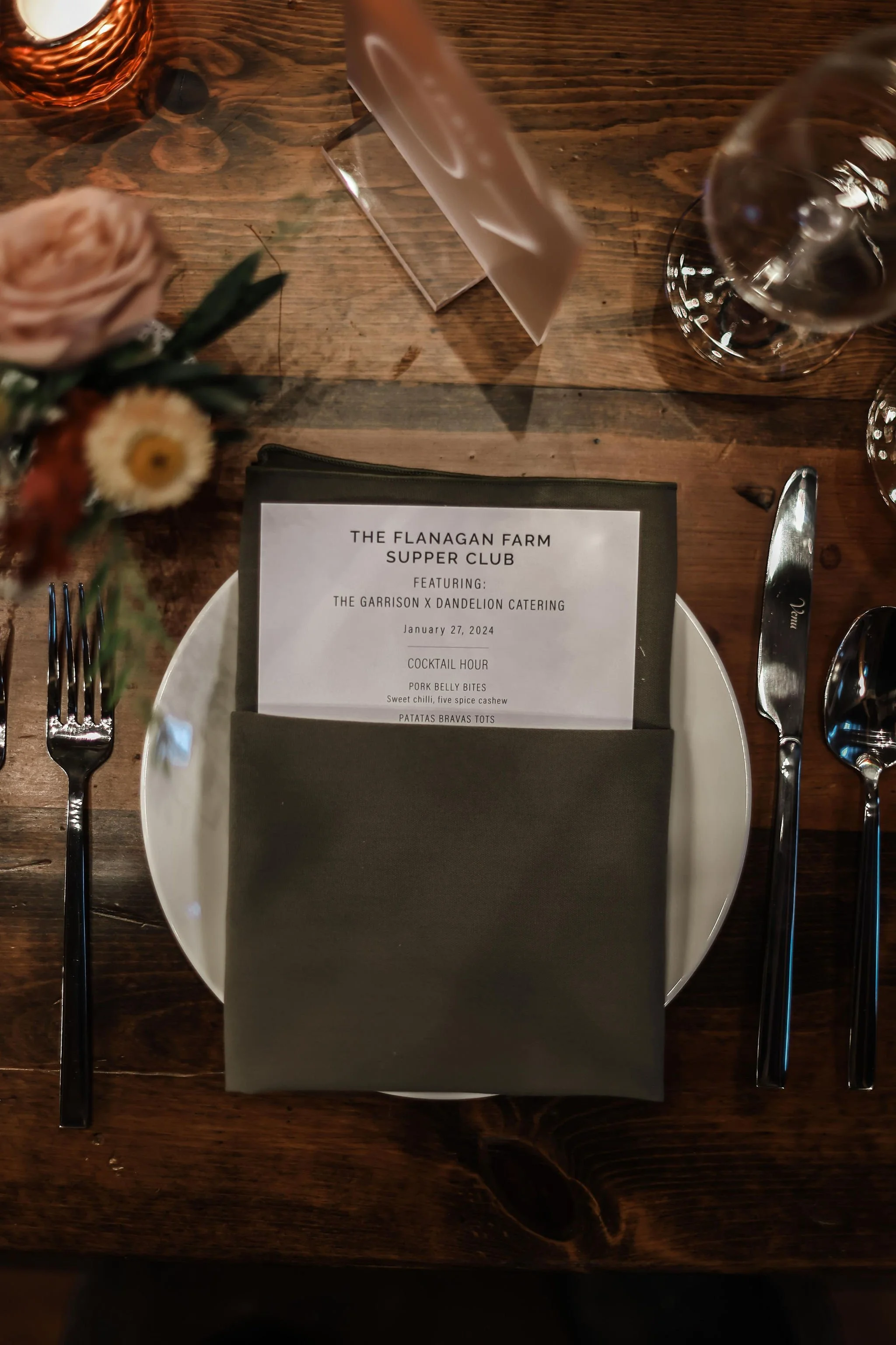 Table setting with a white plate, black napkin, silverware including a fork on the left and a knife on the right, a glass of water, a menu, and a vase of flowers on a wooden table.