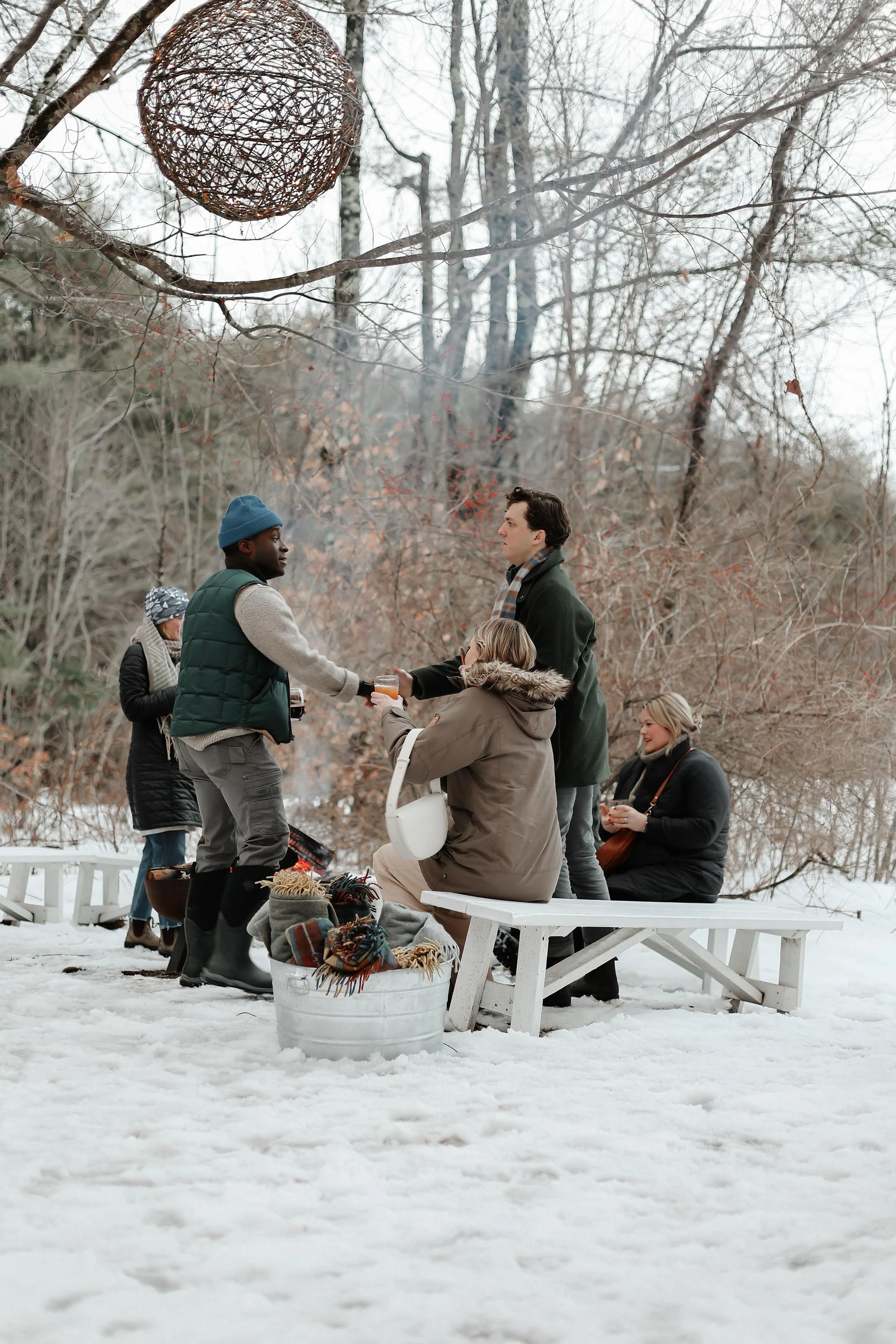 Group of people in winter clothing enjoying a gathering around a fire in a snowy outdoor setting with leafless trees and a wicker lantern hanging from a tree branch.