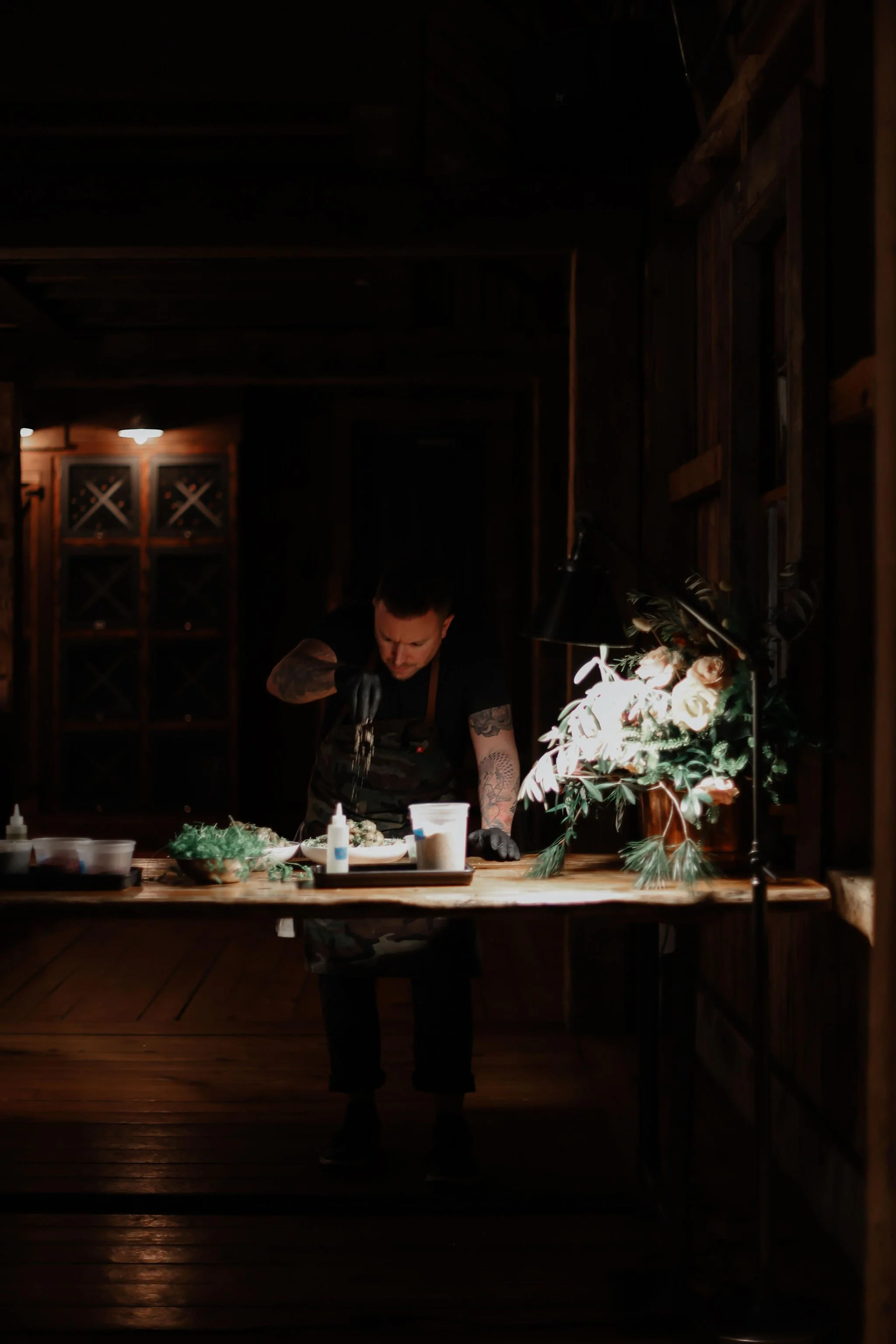 A person working in a dimly lit, rustic kitchen or workshop, preparing food at a wooden counter with various ingredients and bottles.